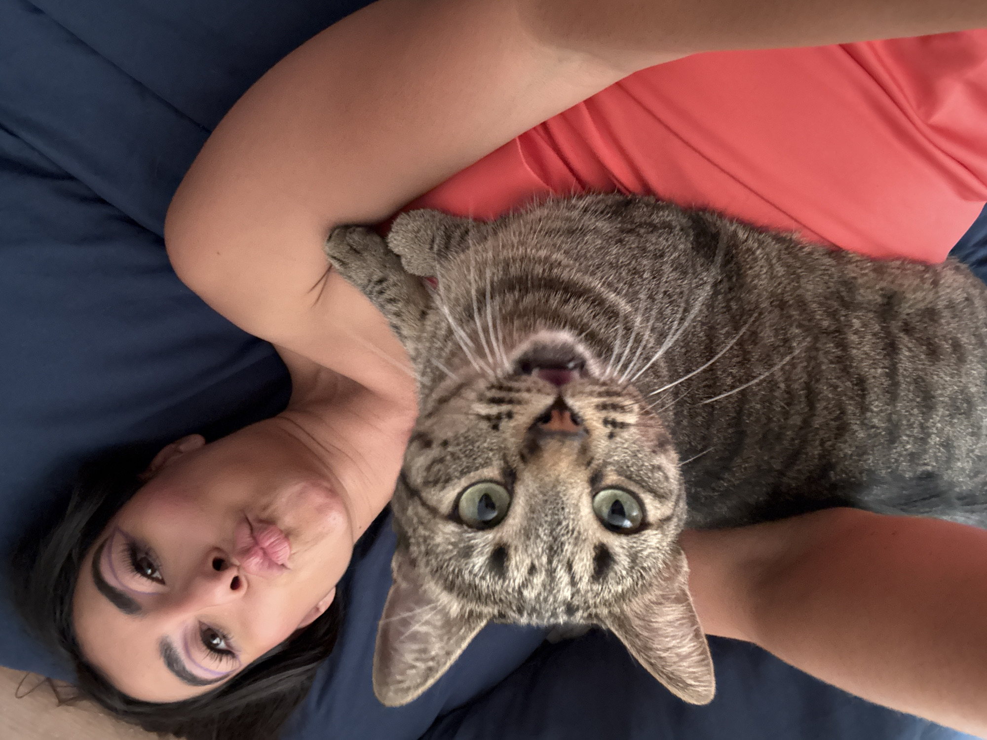 A woman with dark hair and purple eyeliner is lying on a blue surface, possibly a bed or blanket, with a tabby cat on her chest. She is puckering her lips as if posing for a selfie. She is wearing a red shirt. The cat has green eyes and is looking directly at the camera with its mouth slightly open. The photograph appears to have been taken at a tilted angle from directly above. The image does not indicate it was taken in Puebla, Mexico.