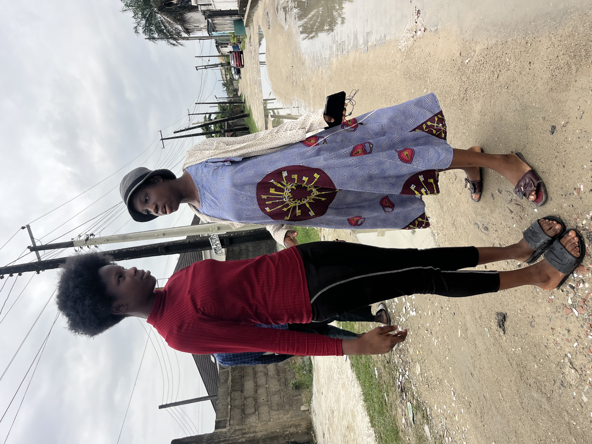 Two young women stand on a dirt road in Ado, Nigeria. The woman on the left wears a red long-sleeved turtleneck and black leggings, with dark, kinky hair. The woman on the right wears a blue and white patterned dress, a light-colored cardigan, and a grey hat. She holds a black smartphone in her right hand. The road is muddy and has large puddles of water. Overhead, power lines crisscross the cloudy sky. In the background, houses and a red car are visible.