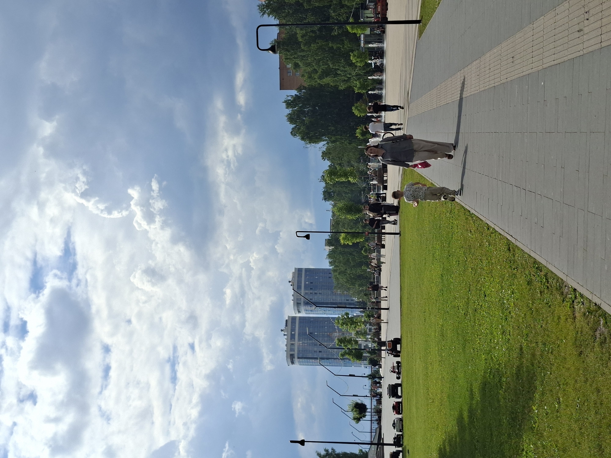 In Izhevsk, Russia, this image captures a public park scene on a partly cloudy day. Two modern, twin high-rise buildings with glass facades dominate the background, suggesting an urban setting. A paved walkway runs through the park, with a neatly mowed green lawn to its left. Several people are walking on the path, including a woman in a grey shirt and beige pants, and a young boy in a patterned shirt and beige shorts walking ahead of her. Further down the path, other individuals are visible, some near fountains, indicating recreational areas. In the foreground on the lawn, several small, ride-on toys resembling cars and tractors are arranged, suggesting a children's play area. The sky is filled with dramatic, textured clouds, with patches of blue sky visible. Streetlights with decorative tops line the walkway.