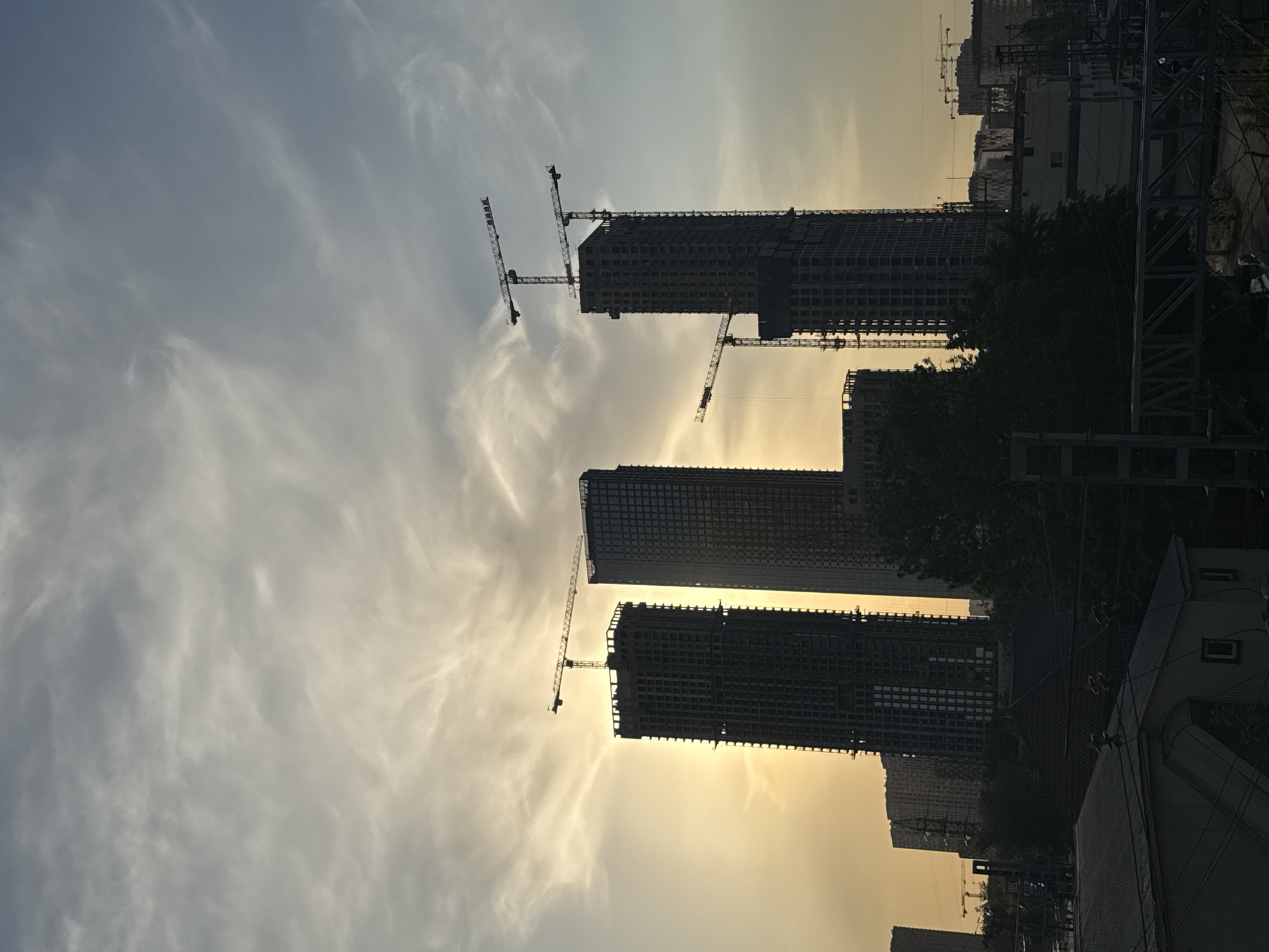 The image depicts a cityscape at dusk or dawn. Several tall buildings are under construction, indicated by the presence of multiple tower cranes. The sky is a mix of gray and yellow, suggesting the sun is either rising or setting. A building, potentially an apartment block or office building, is in the foreground. This suggests that the construction is taking place within or adjacent to an existing urban area. Due to the prompt and image hints, the location is assumed to be in Khimki, Russia. The presence of construction machinery and partially completed structures implies ongoing urban development. There are no people visible in the frame.