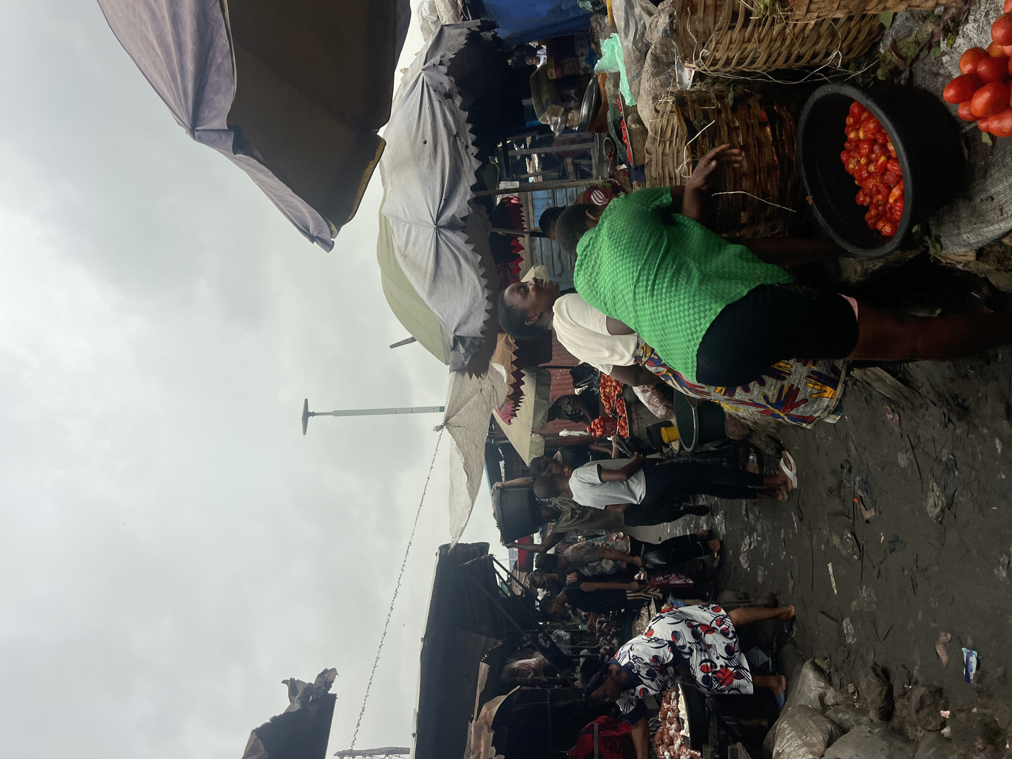 The image depicts a bustling open-air market scene in Nkpor, Nigeria, under an overcast sky. In the foreground, a person wearing a green patterned shirt and black shorts is leaning over a collection of woven baskets, with a black basin filled with red tomatoes to their right. To the left, a person in a white and red floral patterned dress is attending to food items displayed in a large pan. The ground is muddy and littered with debris.
In the midground, several individuals are interacting, with some appearing to be vendors and others customers. One person, wearing a white shirt, stands facing the viewer. Behind them, another person carries a large black basin on their head. A woman in a white shirt and patterned skirt is visible in the center, smiling and looking to her left. Various market stalls with fabric coverings are visible, some appearing weathered.
The background is dominated by the market structures and the cloudy sky. A tall light pole is visible in the distance. The overall atmosphere is one of active commerce in a somewhat unkempt environment.