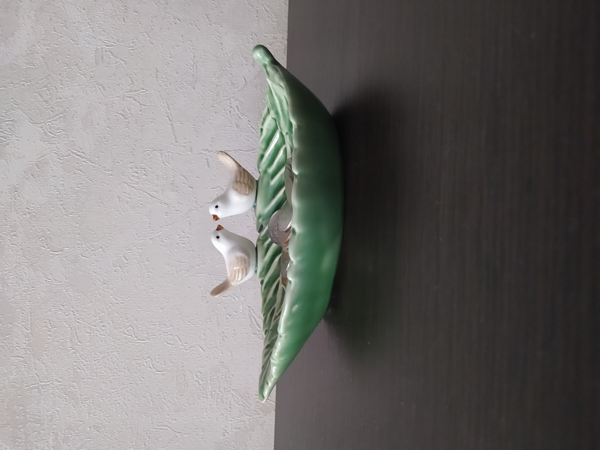 The image shows a decorative ceramic object shaped like a green leaf with two small white birds perched on it. One bird is positioned slightly above and behind the other. The ceramic leaf appears to be holding some coins within its curved form. The object is placed against a textured light-colored wall on the left and a dark wooden surface on the right. The lighting suggests an indoor setting, possibly a home. There is no visible text.