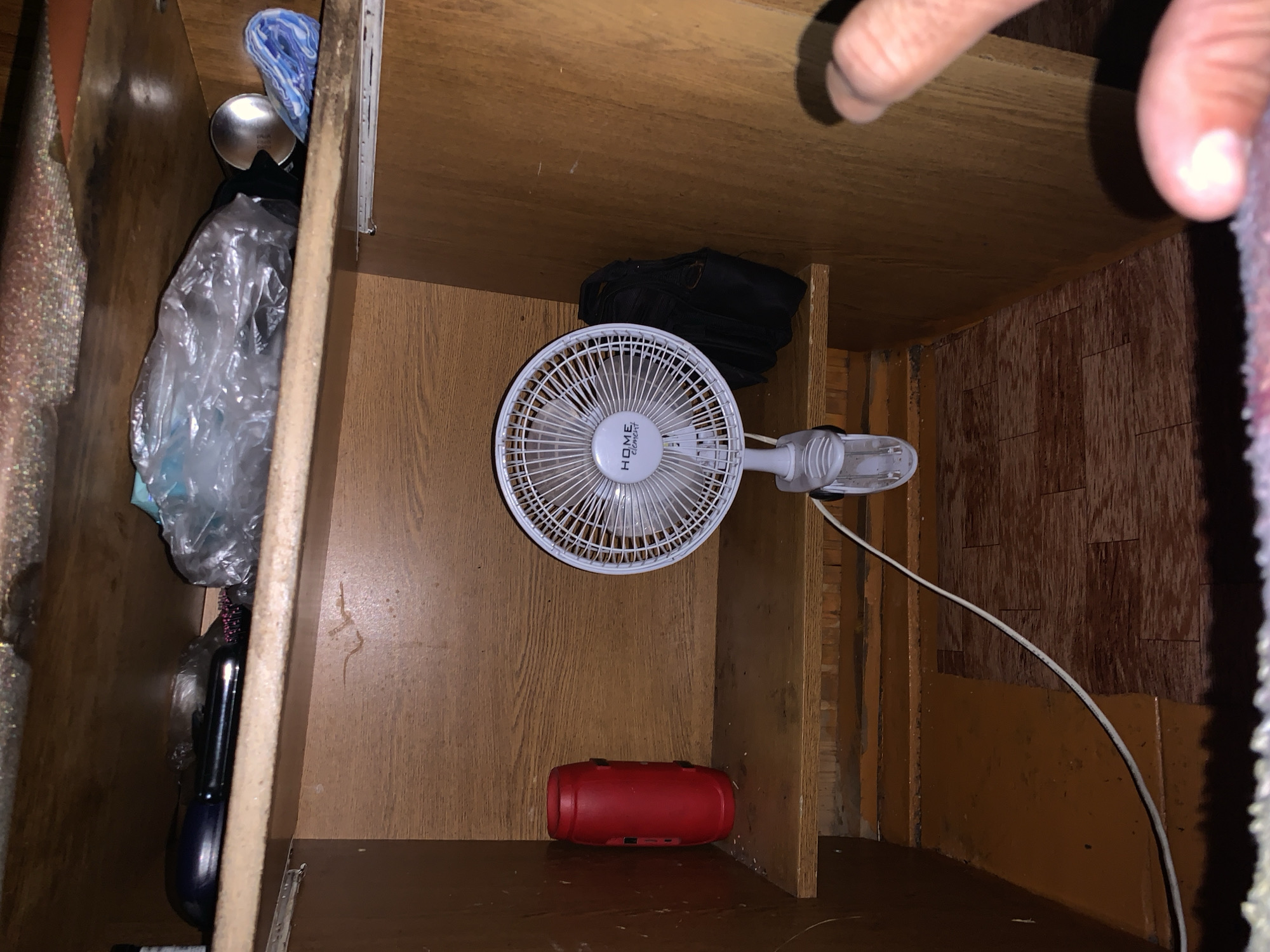 The image depicts the interior of a wooden cabinet, likely within a residential setting in Nizhnee Almurzino, Russia. The cabinet contains several items on its shelves, including a red cylindrical container on the left shelf, and a white electric fan with a black bag behind it on the right shelf. The top shelf contains various indistinct items, some appearing to be in plastic packaging. The environment is dimly lit, and the wooden surfaces show signs of wear. A human hand is visible in the lower right corner, partially obscuring the scene. The overall impression is one of a cluttered, functional storage space.
