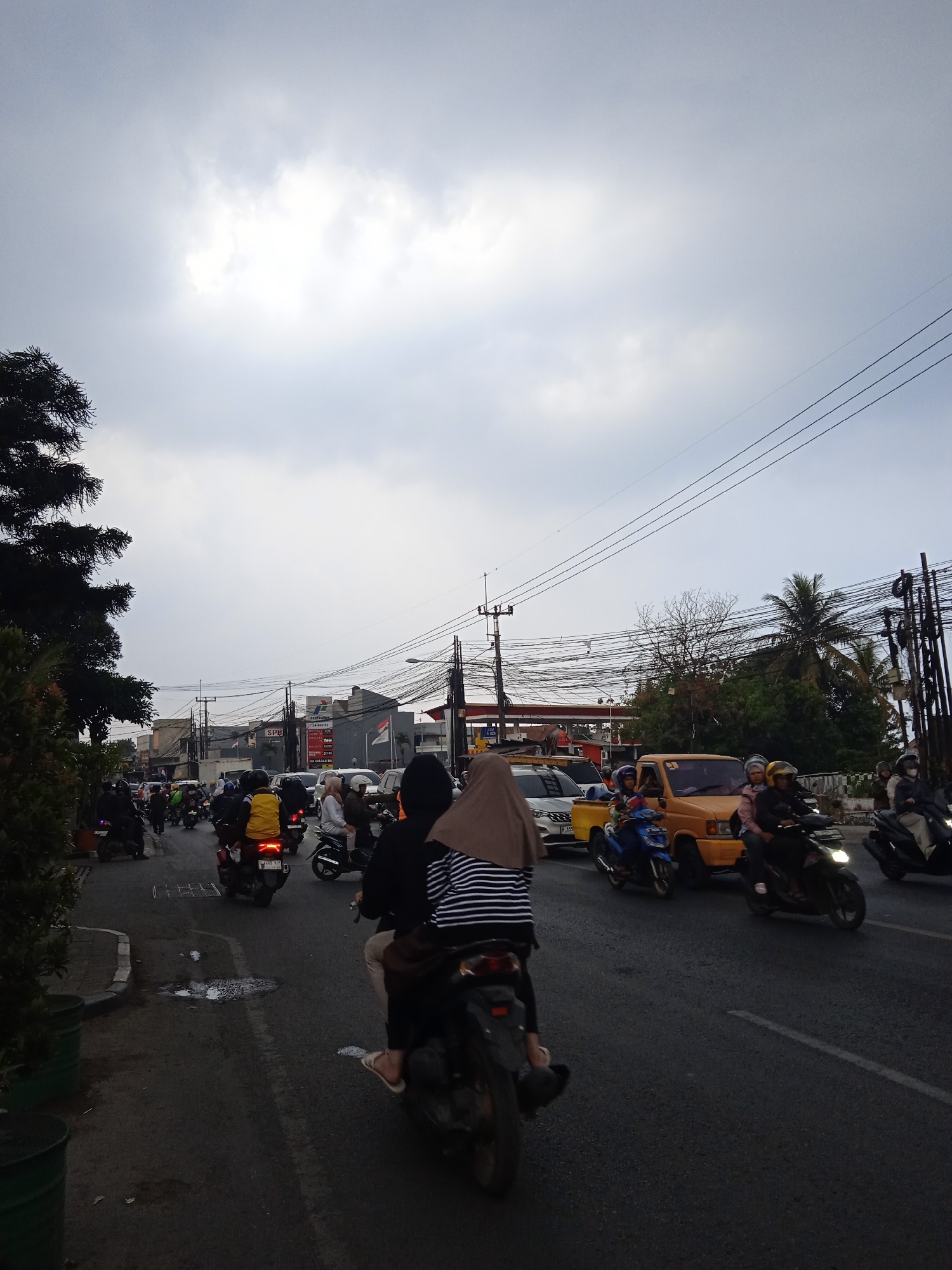 The image depicts a traffic scene in Cileunyi, Indonesia, under an overcast sky. Numerous motorcycles and a few cars are on a paved road, suggesting moderate traffic congestion. Several people are visible riding motorcycles, some with passengers. The environment includes buildings along the roadside, power lines suspended overhead, and some vegetation. A gas station with signage is also present. The overall impression is of a typical urban street scene during daylight hours in Indonesia.