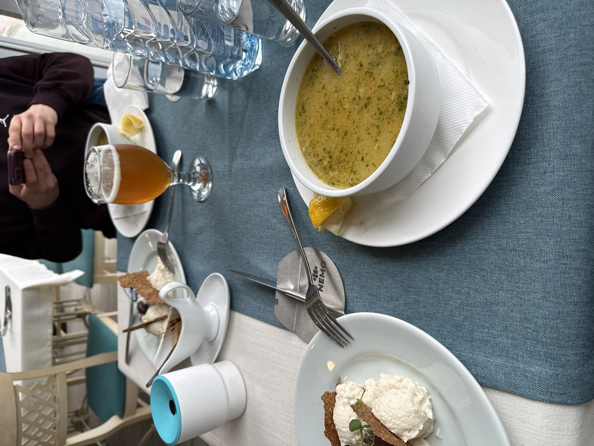 The image depicts a dining table at a restaurant. Soup in a white bowl is placed on a white plate, alongside a lemon wedge. A beer glass filled with beer is visible, with a person in a maroon sweatshirt holding a small bottle to its left. Bottles of water and glasses are arranged near the top of the frame. Below the soup, there is a plate with a dessert and a fork. A white container with a blue top sits next to the dessert. The tablecloth is a muted blue. The scene is likely in a restaurant in Varna, Bulgaria, based on the inscription "Nemo" on a piece of cutlery and associated business information.