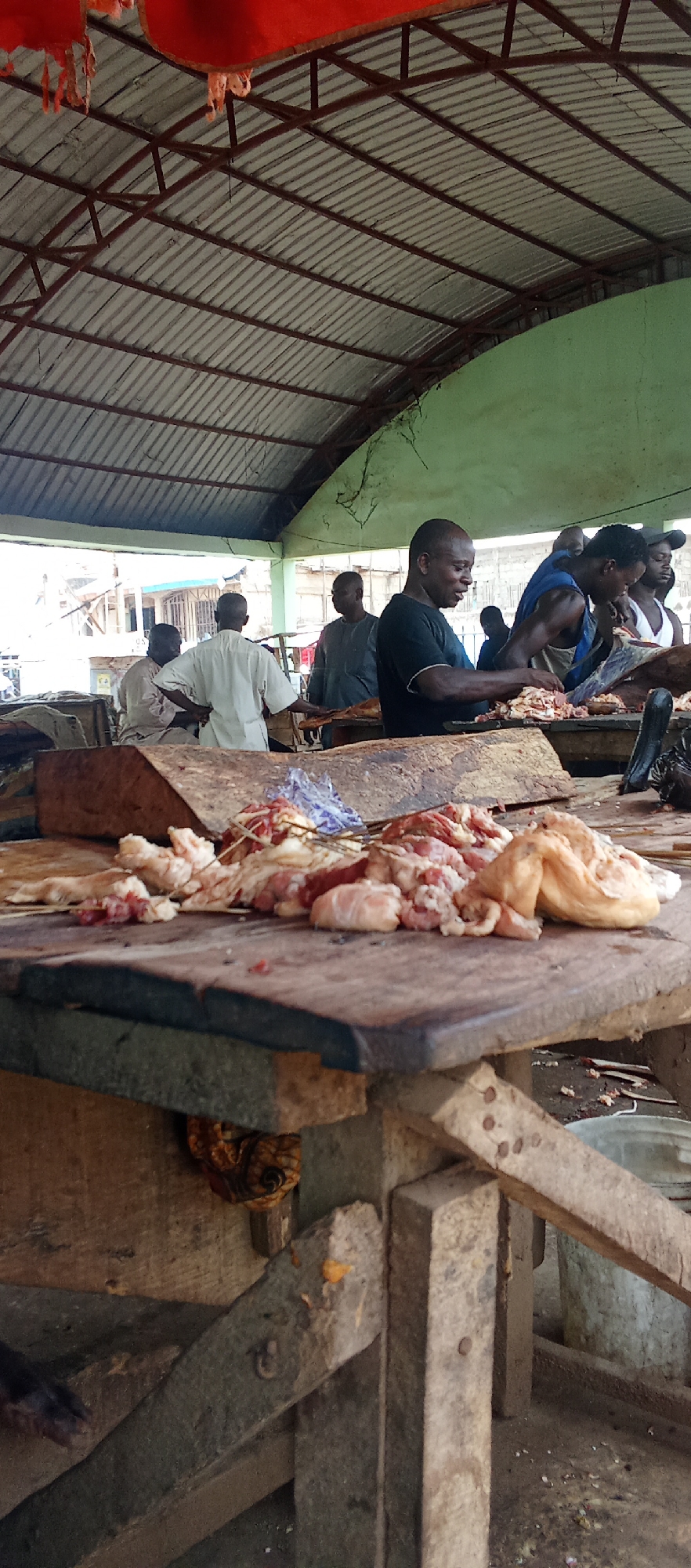 The image depicts a bustling meat market in Akwanga, Nigeria. Under a corrugated metal roof supported by a steel frame, several men are actively butchering and processing meat on a series of crude wooden tables. In the foreground, a table is laden with raw meat, likely freshly cut. Behind it, men stand shoulder-to-shoulder at another table, their hands moving quickly as they work. Other individuals are visible in the background, suggesting a lively commercial environment. A single bucket is also seen in the lower right corner.