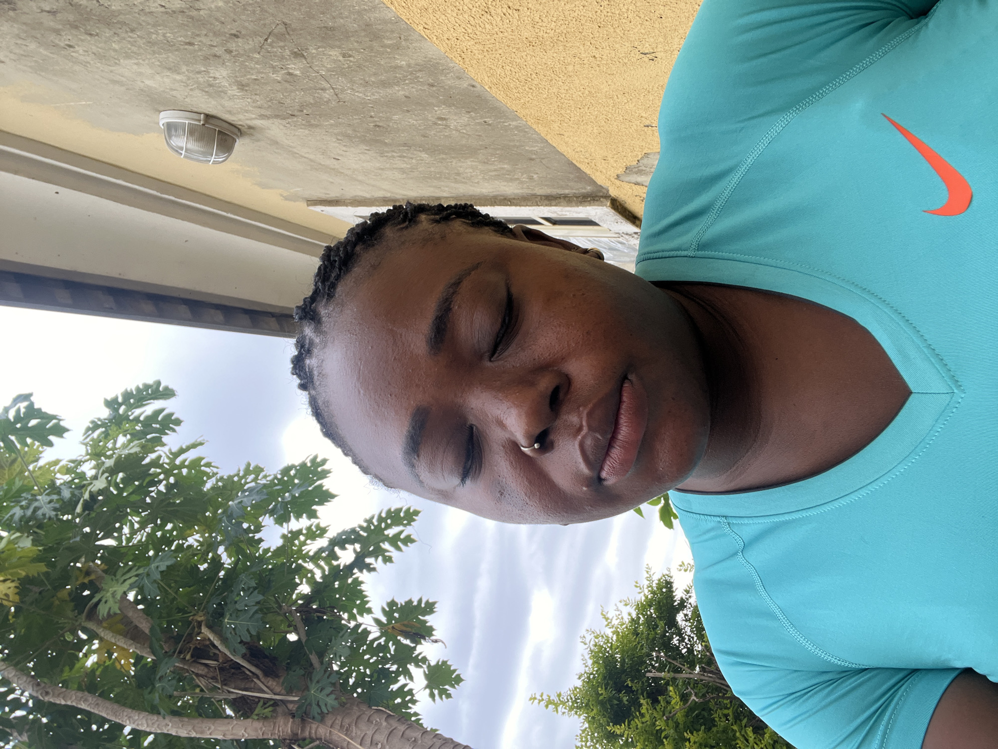 A medium shot shows a Black woman outdoors, her eyes closed and head tilted down. She has braided hair and is wearing a turquoise Nike t-shirt. A papaya tree with large green leaves is visible to her left, and a building with yellow and beige walls and an outdoor light fixture is behind her to the right. The sky is overcast. The location is Abuja, Nigeria.