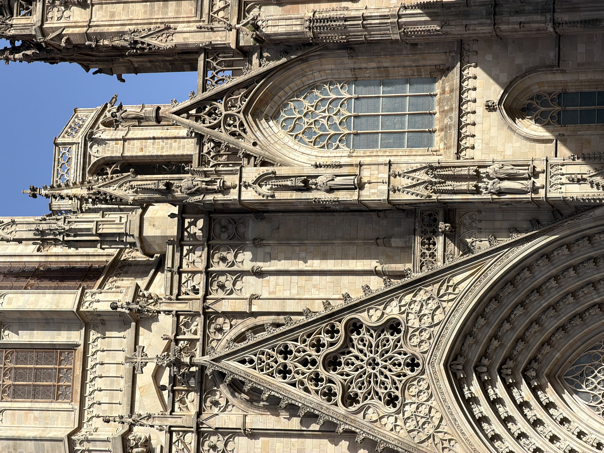 The image shows a close-up, low-angle view of the Gothic Quarter Cathedral in Barcelona, Spain, on a bright, sunny day. The architecture is intricately detailed, featuring stone carvings, ornate windows, statues, and arched structures. The sunlight casts strong shadows, highlighting the textures and depth of the stonework. The blue sky is visible in the upper right portion of the image. There are no people visible in the frame. The scene is static, focusing solely on the majestic facade of the cathedral.