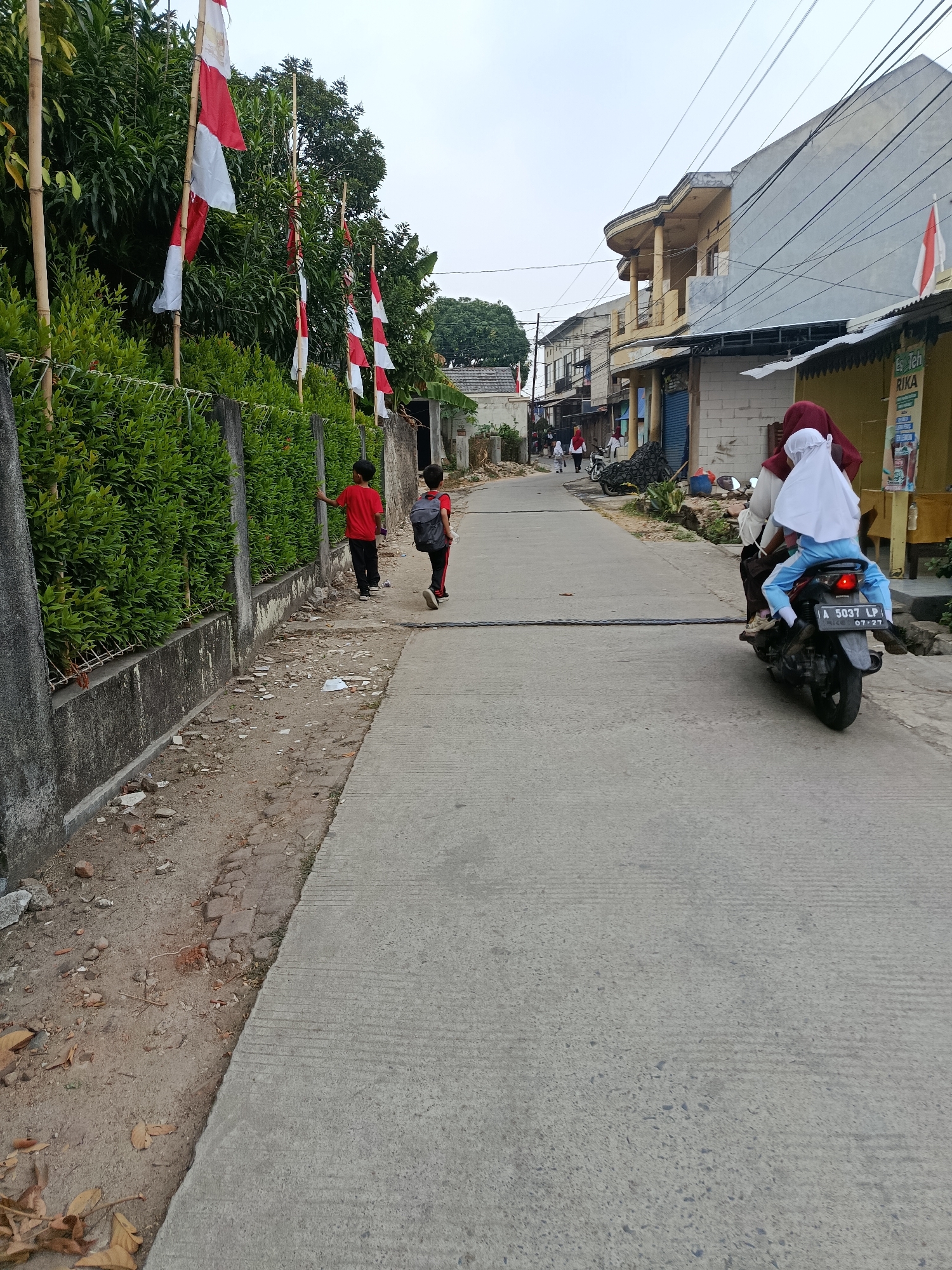 A street scene in Balaraja, Indonesia. The image shows a paved road lined with a concrete wall and a dense green hedge on the left. Indonesian flags are displayed on bamboo poles along the hedge. Several children are walking on the side of the road, and a motorcycle carrying two individuals is moving away from the viewer. Buildings are visible on the right side of the street, with one structure featuring a distinctive curved balcony. The sky is overcast.