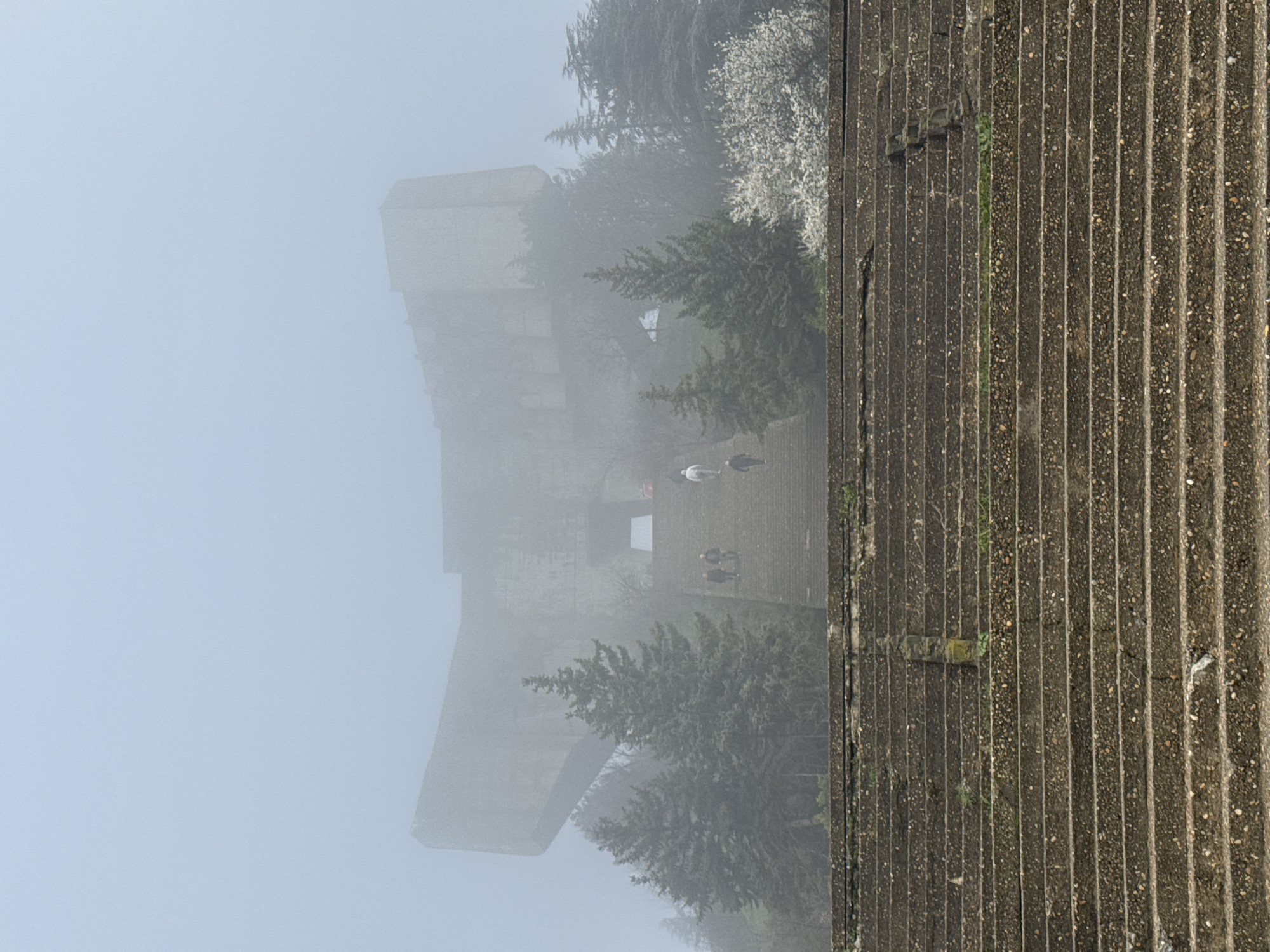 The image depicts a concrete structure, likely part of the Monument to the Bulgarian-Soviet Friendship near Varna, Bulgaria, shrouded in fog. Several people are visible near the base of the monument. The concrete facade shows vertical striations with visible wear and vegetation. The monument fades into a dense fog. Trees and shrubbery are present, partially obscured by the fog.