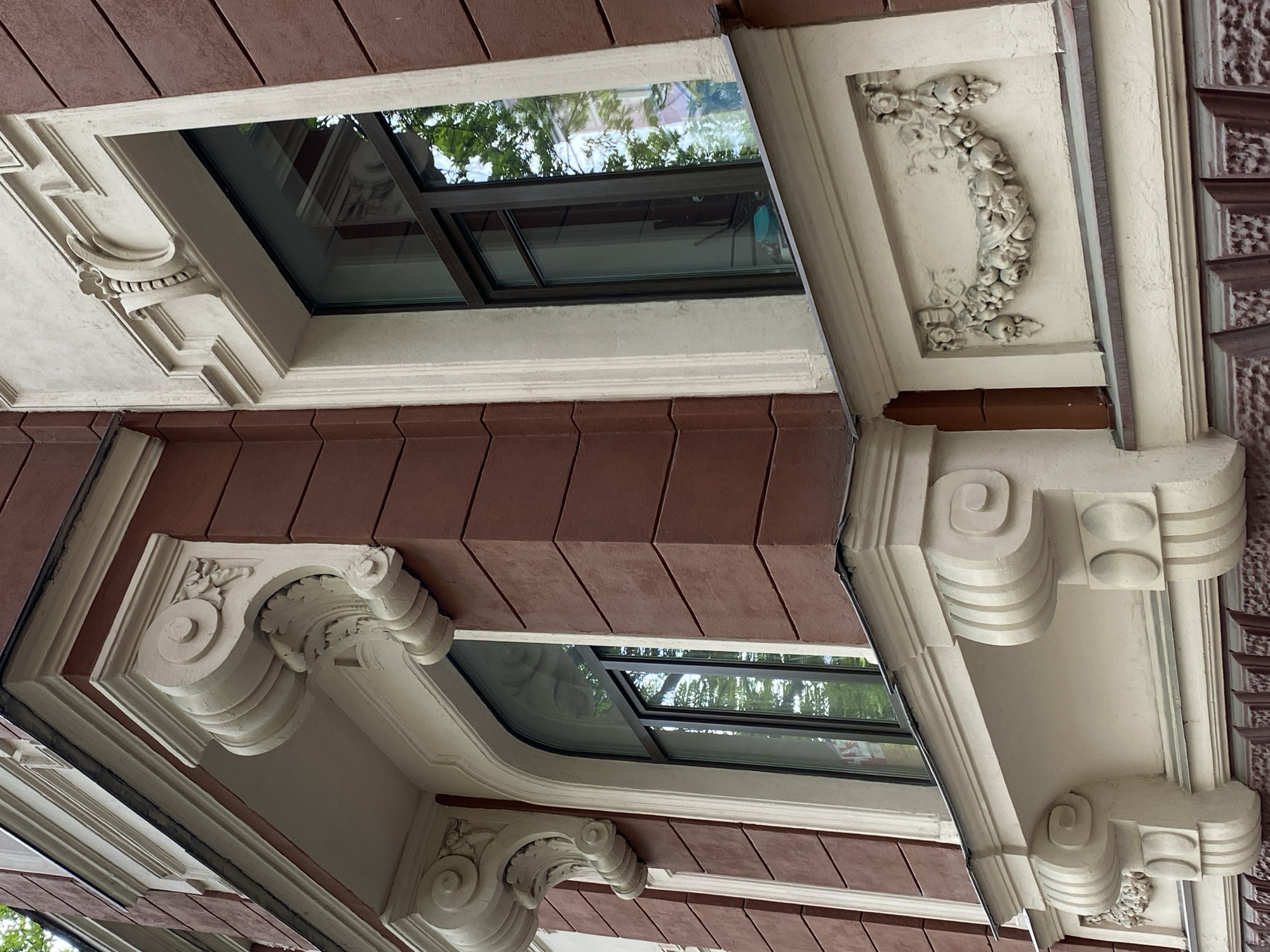 The image displays a close-up, upward-angled view of an ornate building facade, showcasing intricate architectural details. The primary subjects are decorative window surrounds and structural corbels, set against a background of reddish-brown siding.
Several windows are visible, appearing to be skylights or dormer windows, framed by thick, light-colored stucco or plasterwork. These frames feature elaborate moldings, often incorporating decorative elements such as scrolls and relief patterns. The window panes reflect bright daylight and the green foliage of trees, indicating an outdoor setting during the day.
Below and around the windows, a variety of ornate corbels or brackets are present. Some exhibit classical design elements like volutes and acanthus leaves, suggesting a blend of styles. One prominent detail is a carved floral and fruit garland, possibly made of stucco, adorning a recessed panel. Other brackets feature multiple curvilinear scrolls and rounded forms, with some even incorporating more geometric patterns.
The building's main surface is covered in dark reddish-brown material, possibly tiles or shingles, arranged in horizontal rows with a slight overlap, providing a textured contrast to the smooth, pale stucco. The overall architectural style, with its rich detailing and stucco work, is consistent with historic buildings found in Saint Petersburg, Russia. No people or specific events are depicted, and no visible text is present.