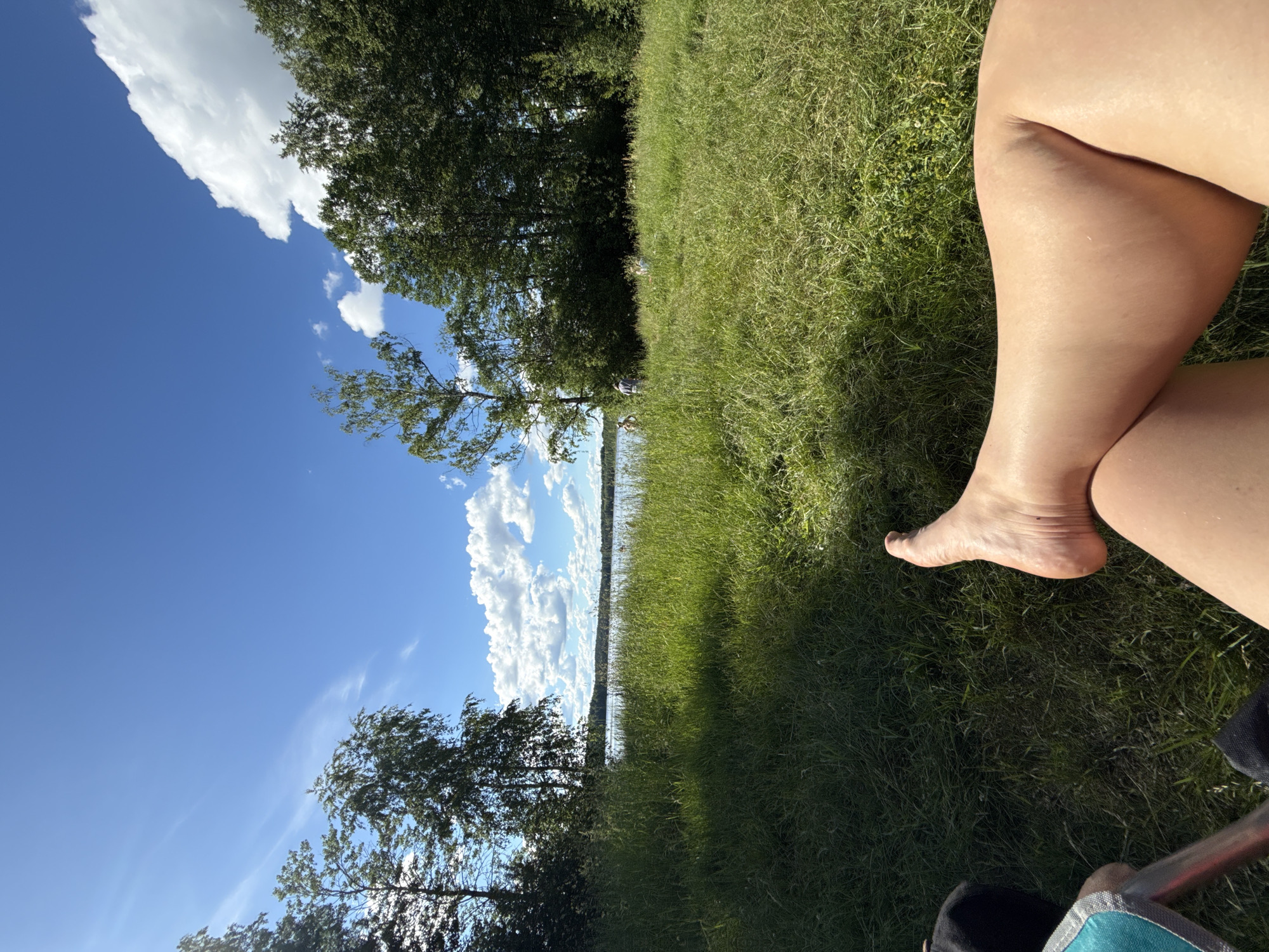 The image captures a sunny day with a clear blue sky dotted with fluffy white clouds. A person's bare legs and feet are in the foreground, resting on a patch of green grass. Beyond the grass, a tranquil lake is visible, bordered by a dense line of trees. The scene suggests a peaceful outdoor setting, possibly a park or a lakeside recreational area in or near Korotsko, Russia. There are a few distant figures in the middle ground, appearing to be enjoying the outdoors by the water.