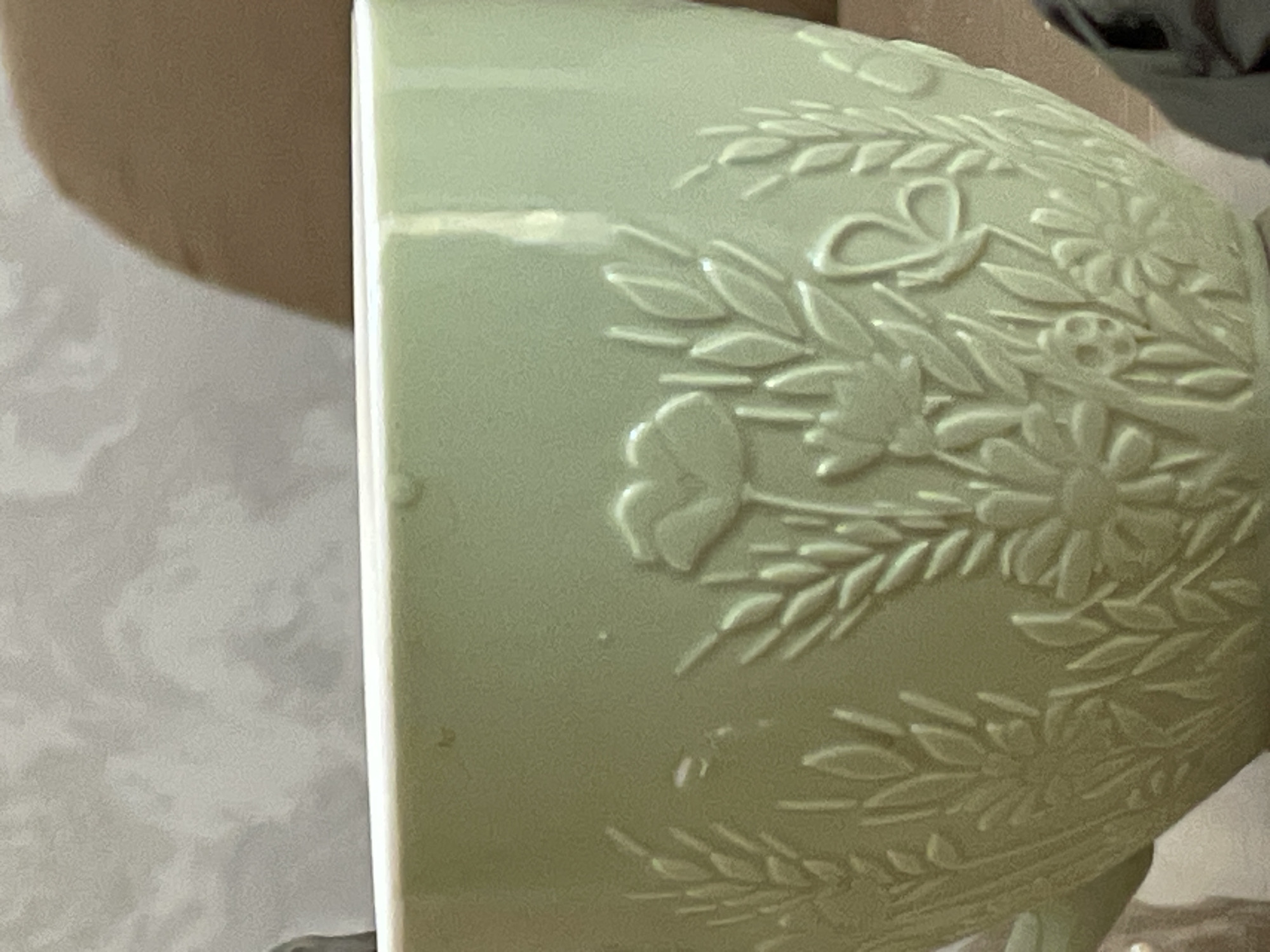 A close-up, eye-level shot focuses on a light green ceramic cup with embossed floral and wheat designs. The cup has a smooth, matte finish and a white rim. The intricate relief features wheat stalks, daisies, poppies, and a butterfly. The background is blurred, showing a textured wall and a brown object. The lighting is soft and diffused. The image is cropped, with the bottom portion of the cup and a glimpse of a dark object on the right edge visible. The scene does not contain any identifiable landmarks or elements that would indicate its location in Tashkent, Uzbekistan.
