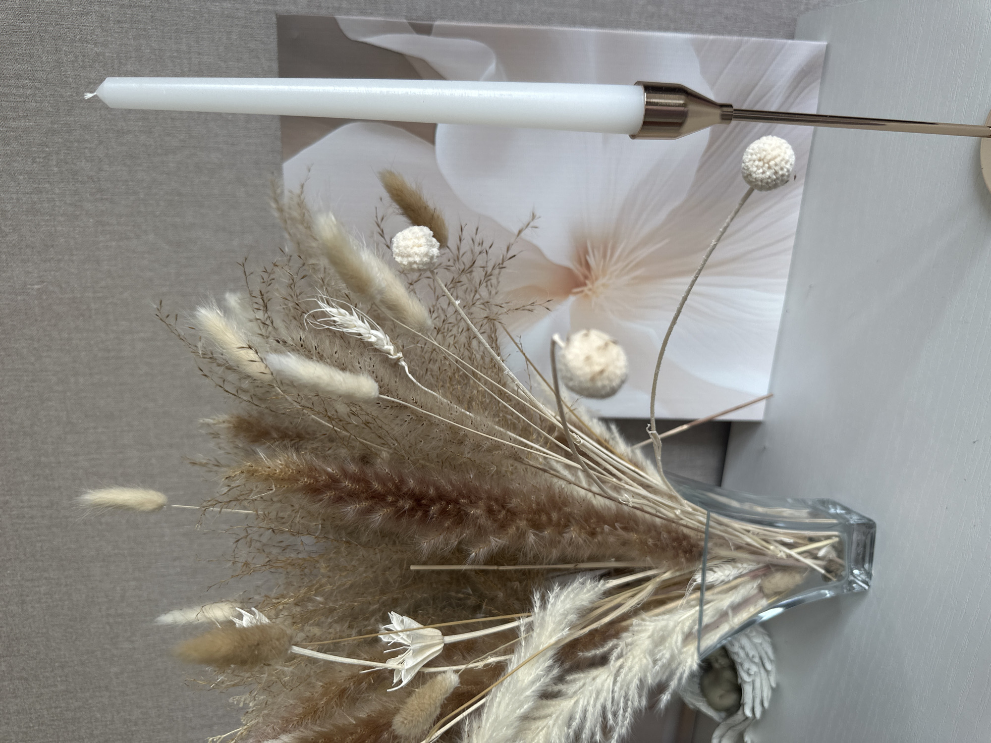 A white taper candle stands in a gold candle holder to the right of a glass vase filled with dried floral arrangements. The dried arrangement features pampas grass, bunny tails, and other beige and white fluffy botanicals. Behind the vase and candle is a canvas print of a large, light pink flower. The scene is set against a textured grey wall and sits on a white surface. The location is not specified in the image.