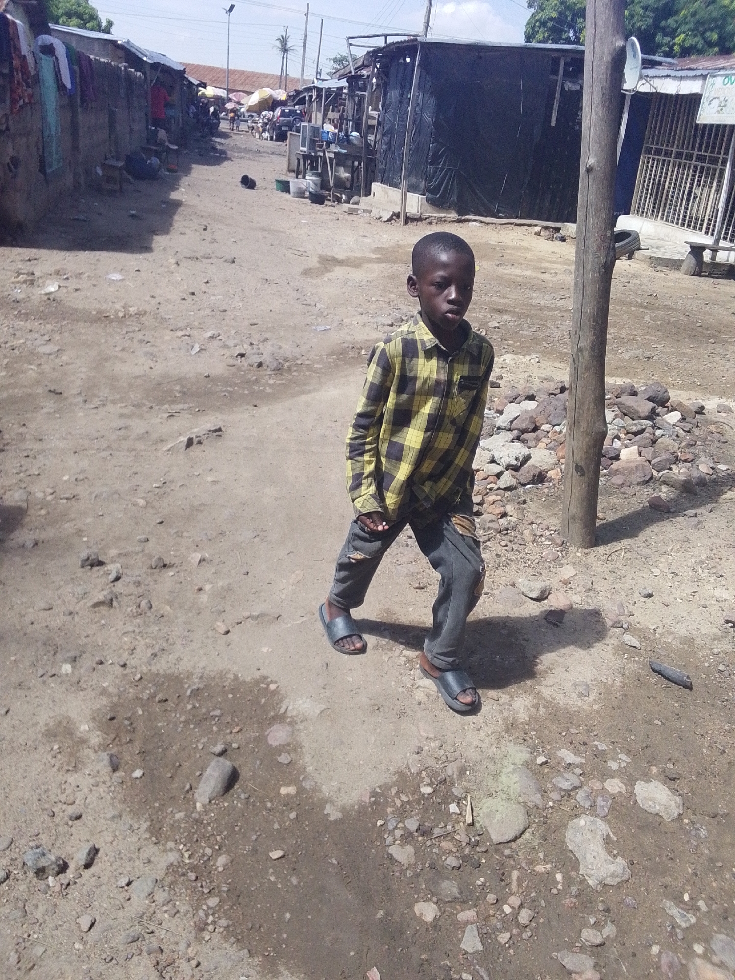 A young boy stands on a dirt road in Lokoja, Nigeria. The boy is wearing a yellow and black plaid shirt and gray jeans with a hole in the knee. He wears sandals on his feet. He is standing next to a wooden pole. The dirt road is uneven and has small rocks scattered across it. In the background, there are buildings and people. There are clothes hanging out to dry on the building to the left.