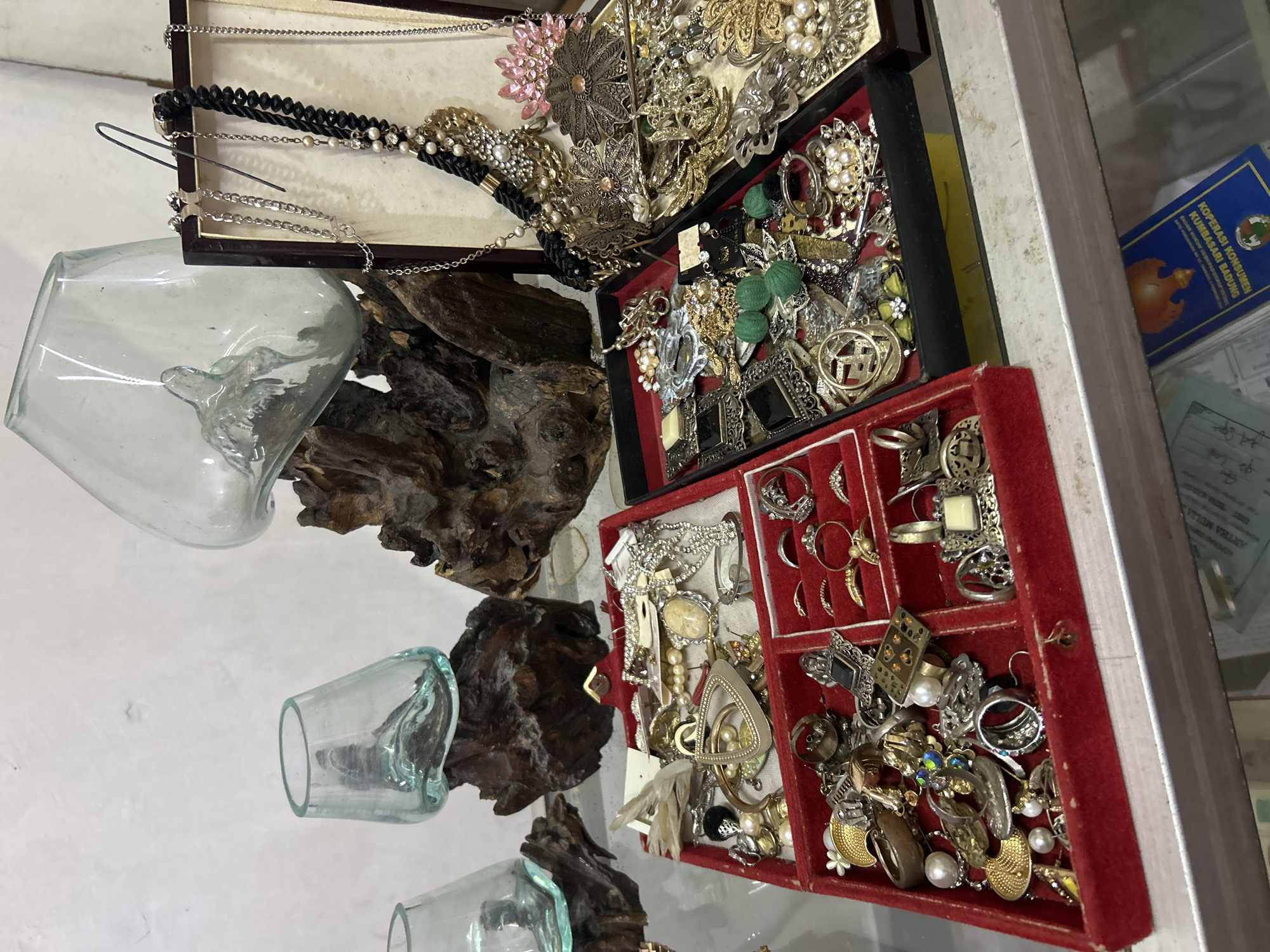 In a marketplace stall in Denpasar, Indonesia, an assortment of jewelry is displayed on a shelf. The jewelry is organized in several red velvet-lined trays and a dark-colored box. Items include necklaces, rings, brooches, and earrings, featuring various metals, pearls, and gemstones. Behind the jewelry, several clear glass vessels with organic, undulating shapes are precariously balanced on pieces of dark, gnarled driftwood. The background is a plain, light-colored wall, suggesting an indoor setting or a covered stall.