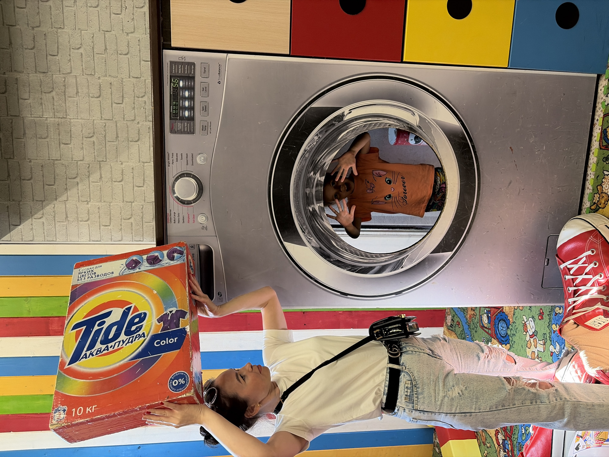 A woman is standing in front of a giant washing machine prop in Izhevsk, Russia, holding a large box of Tide detergent over her head. The prop washing machine has a child inside, peeking out from the drum. The background features a colorful striped wall and a brick-patterned wall. The floor is covered with a rug featuring cartoon animals and vehicles. The woman is wearing a white t-shirt, ripped jeans, and a black crossbody bag. The child is wearing an orange t-shirt with a cat print.