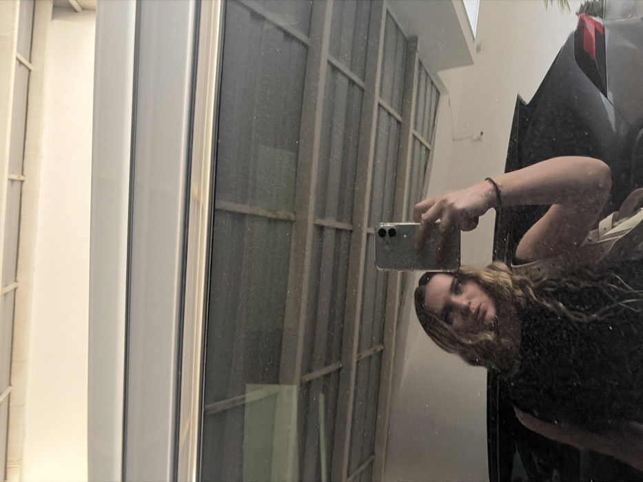The image depicts a female subject captured in a selfie-style reflection within a large, rectangular reflective surface, located in San Bernardino Tlaxcalancingo, Mexico. The subject, a woman with long, wavy blonde hair and wearing a dark, sleeveless top, is positioned in the lower-left quadrant of the frame. Her right arm is raised, holding a light-colored smartphone with a dual-camera setup, angled to capture her reflection. A dark bracelet is visible on her right wrist, and a light-colored strap, possibly from a bag, is visible over her left shoulder. Her gaze is directed towards the camera lens.
The background visible in the reflection consists of a multi-paneled, light-colored overhead structure, indicative of a ceiling or covered area, possibly a garage or carport. Below this structure and behind the woman, the dark bodywork of a vehicle, appearing to be an SUV or crossover, is partially reflected, with a section of its rear or side visible on the right. The reflective surface itself shows minor dust specks and streaks.