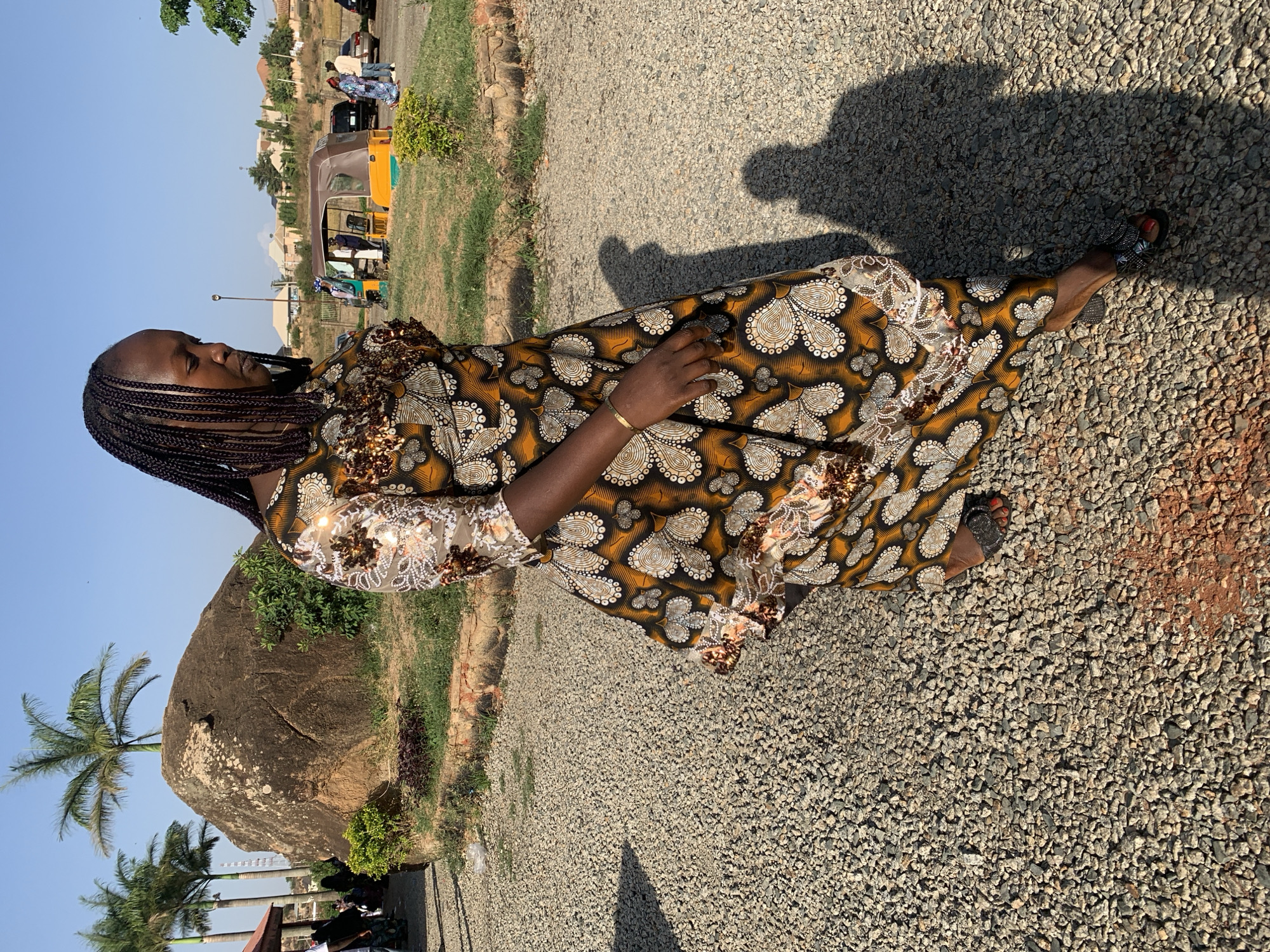 A full shot captures a woman in a vibrant, flowing dress standing on a gravel surface. The dress features a mix of brown and beige with a paisley-like pattern. She has dark hair styled in braids and is wearing a gold bracelet on her left wrist. Her left hand is resting on her dress.
Behind her, there is a large, smooth rock formation and a few palm trees. Further in the background, a yellow auto-rickshaw (likely a "Keke Napep" as they're called in Nigeria) and other vehicles are visible, along with buildings suggesting an urban area. The sky is clear and blue.
The scene likely takes place in Jos, Nigeria.