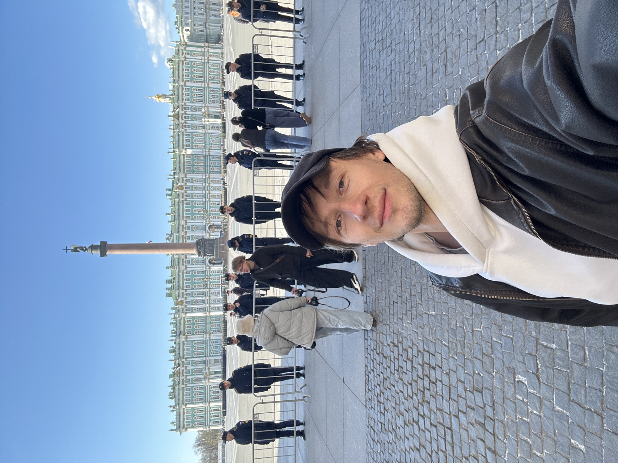 The image is a selfie, rotated 90 degrees clockwise, featuring a man in the foreground against the backdrop of Palace Square in Saint Petersburg, Russia. The man wears a black baseball cap, a white hoodie, and a black leather jacket, looking towards the camera with a slight smile.
Behind him, under a clear blue sky with a few white clouds, stands the majestic Winter Palace, a large building with light green and white facades and a distant golden dome or spire. To the left, the prominent Alexander Column, topped with a statue, rises from the square.
The foreground and mid-ground consist of a wide plaza paved with grey cobblestones. Numerous people are visible, many arranged in lines behind metal barriers. A significant number appear to be uniformed security or military personnel in dark clothing and hats. Among the civilians, one individual in a light grey puffer jacket is seen holding a camera.
The scene depicts a daytime setting, bright and sunny. The presence of barriers and uniformed individuals suggests an organized event, a public gathering, or a queue for a major tourist attraction. No visible text is discernible in the image.