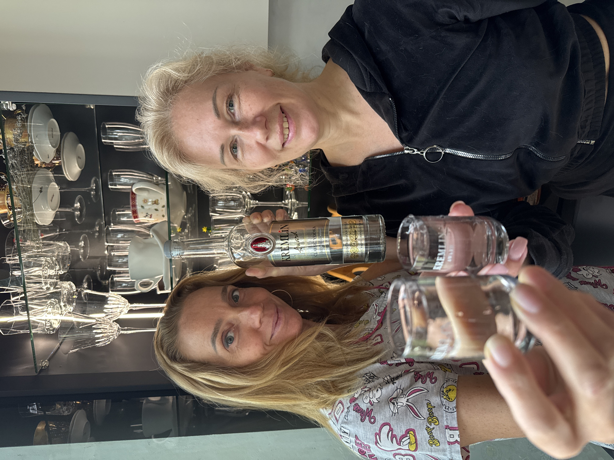 Two Caucasian women are posing for a photograph indoors. The woman on the top is wearing a black velour tracksuit. The woman on the bottom has long blonde hair and is wearing a gray pajama top with cartoon characters. They are each holding up a shot glass and a bottle of Kremlin Award Classic Vodka. Behind them is a glass-fronted cabinet containing dishes and glassware. The image does not contain enough information to identify the location in Steklyannyi, Russia.