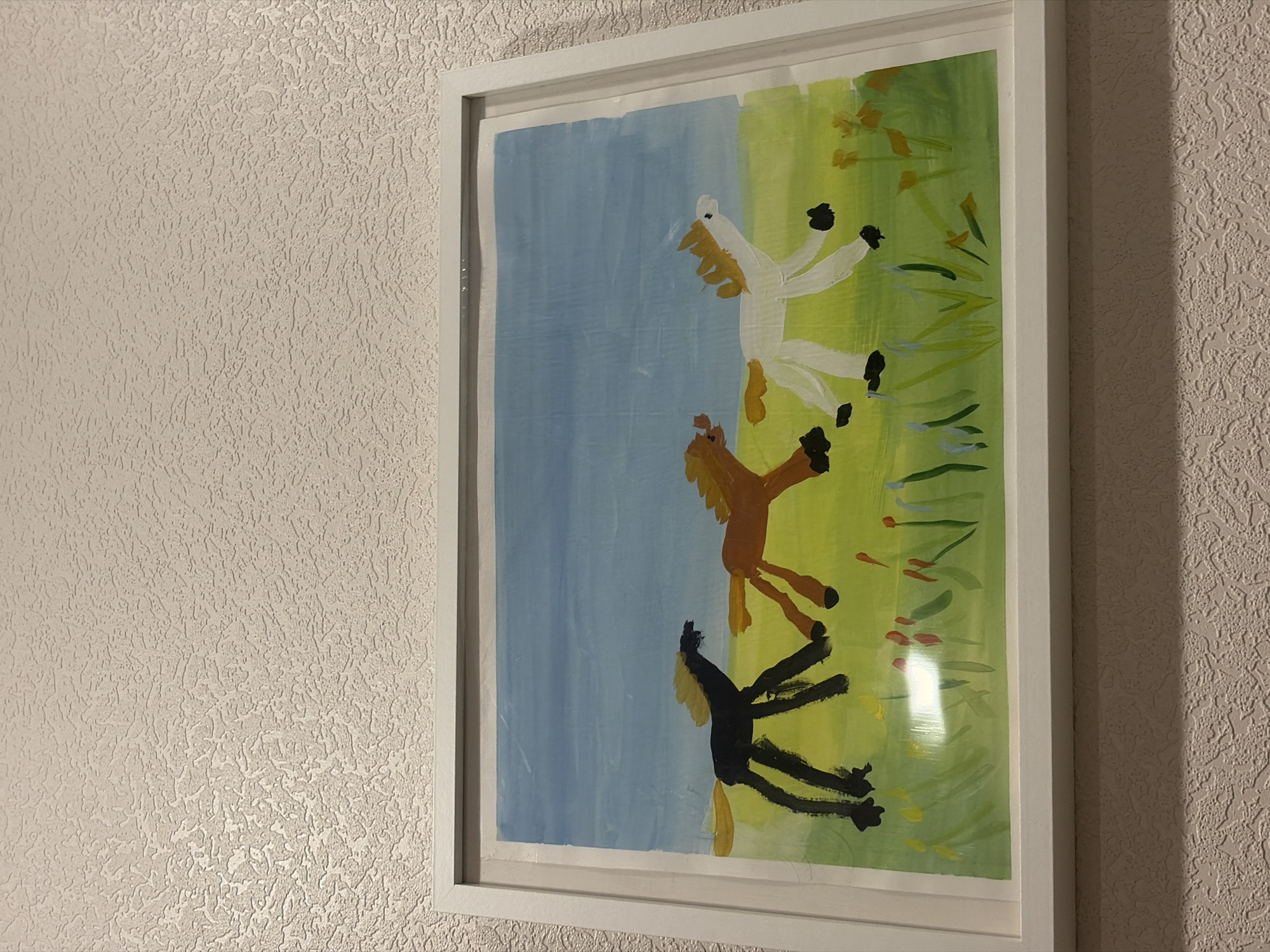The image depicts a framed child's painting of three horses, hung on a textured wall. The painting is roughly divided into two sections: a blue area on the left, and a green area on the right. Three horses are painted, a white horse at the top, a brown horse in the middle, and a black horse at the bottom. The frame appears to be white. The location is not Saint Petersburg, Russia.