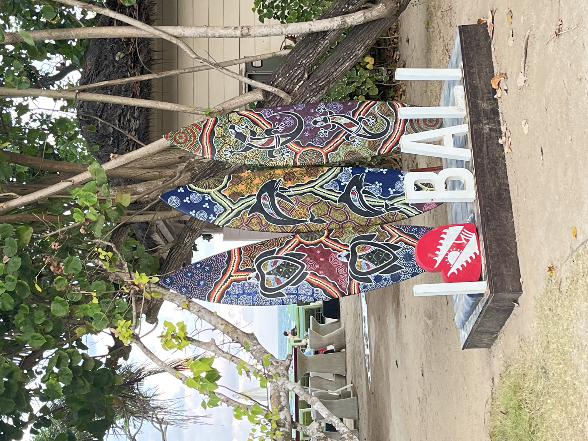 In Kuta Selatan, Indonesia, three vibrantly painted surfboards stand upright on a sandy beach, leaning against tree trunks. The surfboards feature intricate dot-art designs with motifs of sea turtles, dolphins, and lizards in shades of blue, red, yellow, green, and purple. In front of the surfboards, large white letters spell out "BALI" on a wooden platform. A red, heart-shaped sculpture with white designs sits to the left of the letters. The background reveals more trees, a thatched-roof structure, and a glimpse of the ocean with a person in the water. The scene is set on a beach with sparse vegetation and scattered dry leaves on the sand.