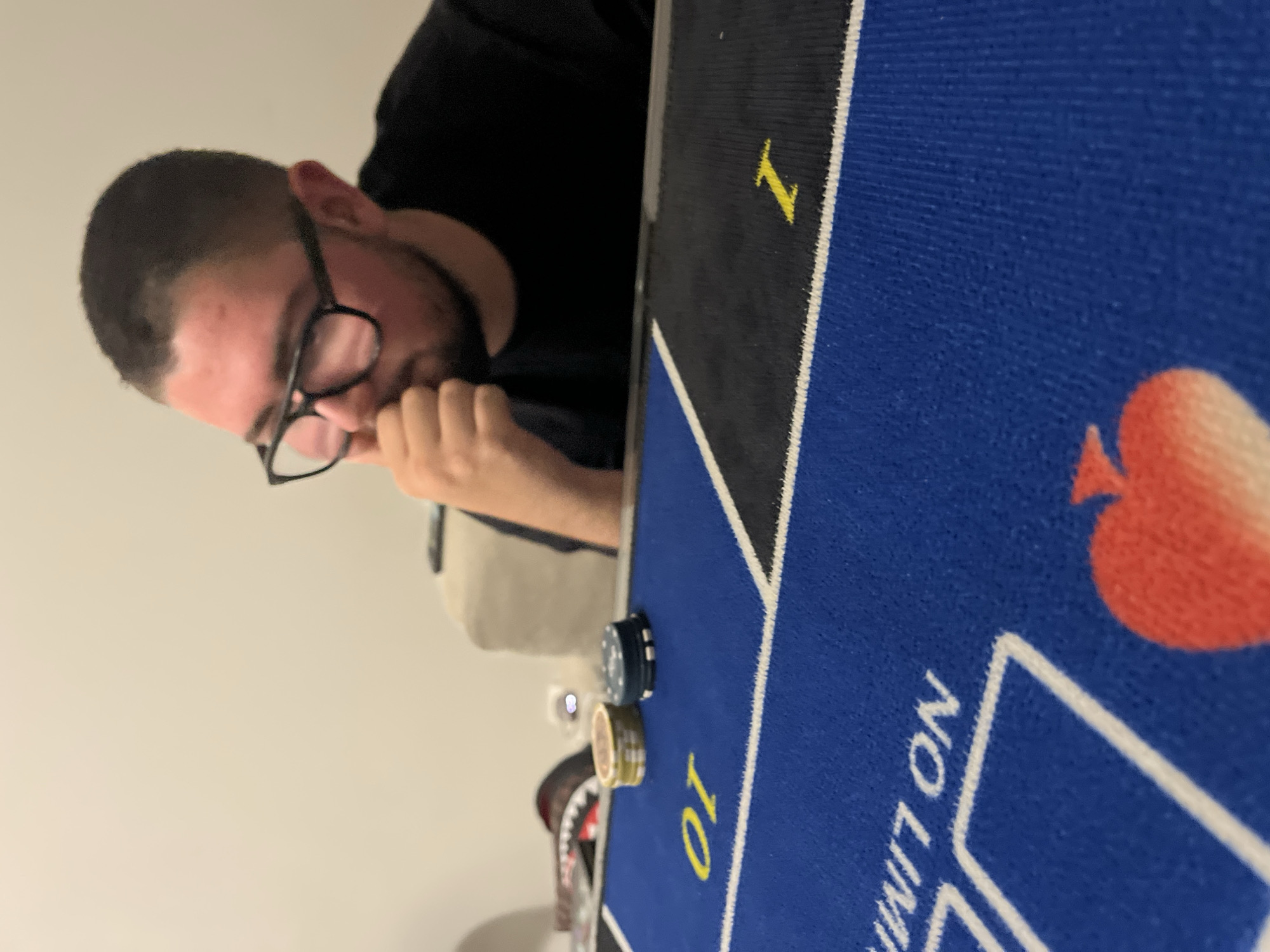 A man with short dark hair and glasses is sitting at a poker table. He is wearing a black shirt and has a beard. He appears to be contemplating a decision, as his hand is on his chin. The poker table has a blue felt surface with white lines and yellow lettering that reads "NO LIMIT" and "10". There is also a red heart symbol on the table. There are poker chips stacked near the edge of the table. The background is a plain light-colored wall. I cannot determine the location of the image in Burgas, Bulgaria.