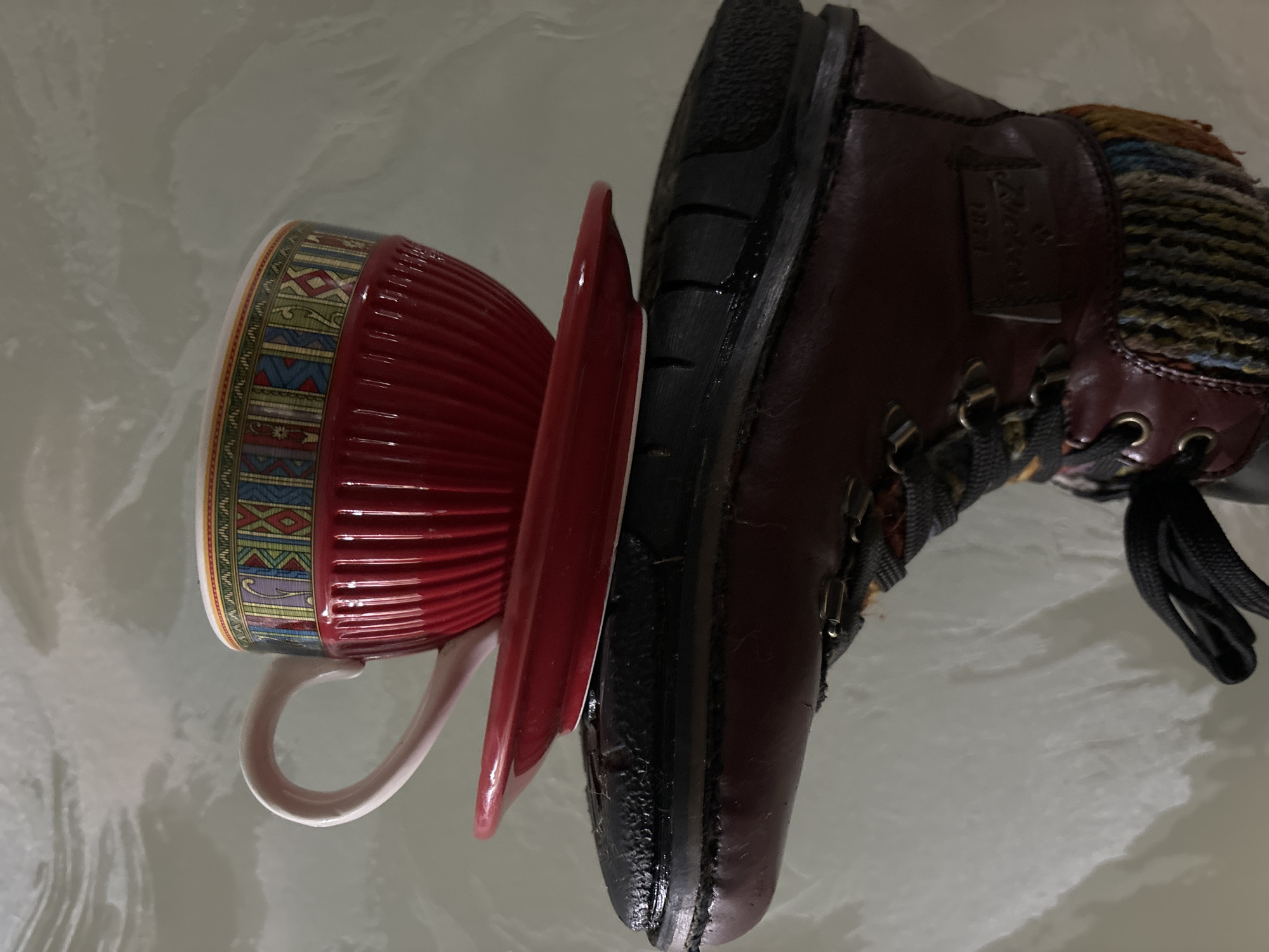 A close-up shot depicts a boot crushing a plate and a mug on a reflective surface. The boot is brown leather with black laces and a black sole. The plate is red, and the mug is ridged with red and decorative multicolored patterns on the rim. The reflective surface is light gray, possibly glass or polished stone. The location is not discernible from the image. The city of Chaikovsky, Russia, is not identifiable from the image.