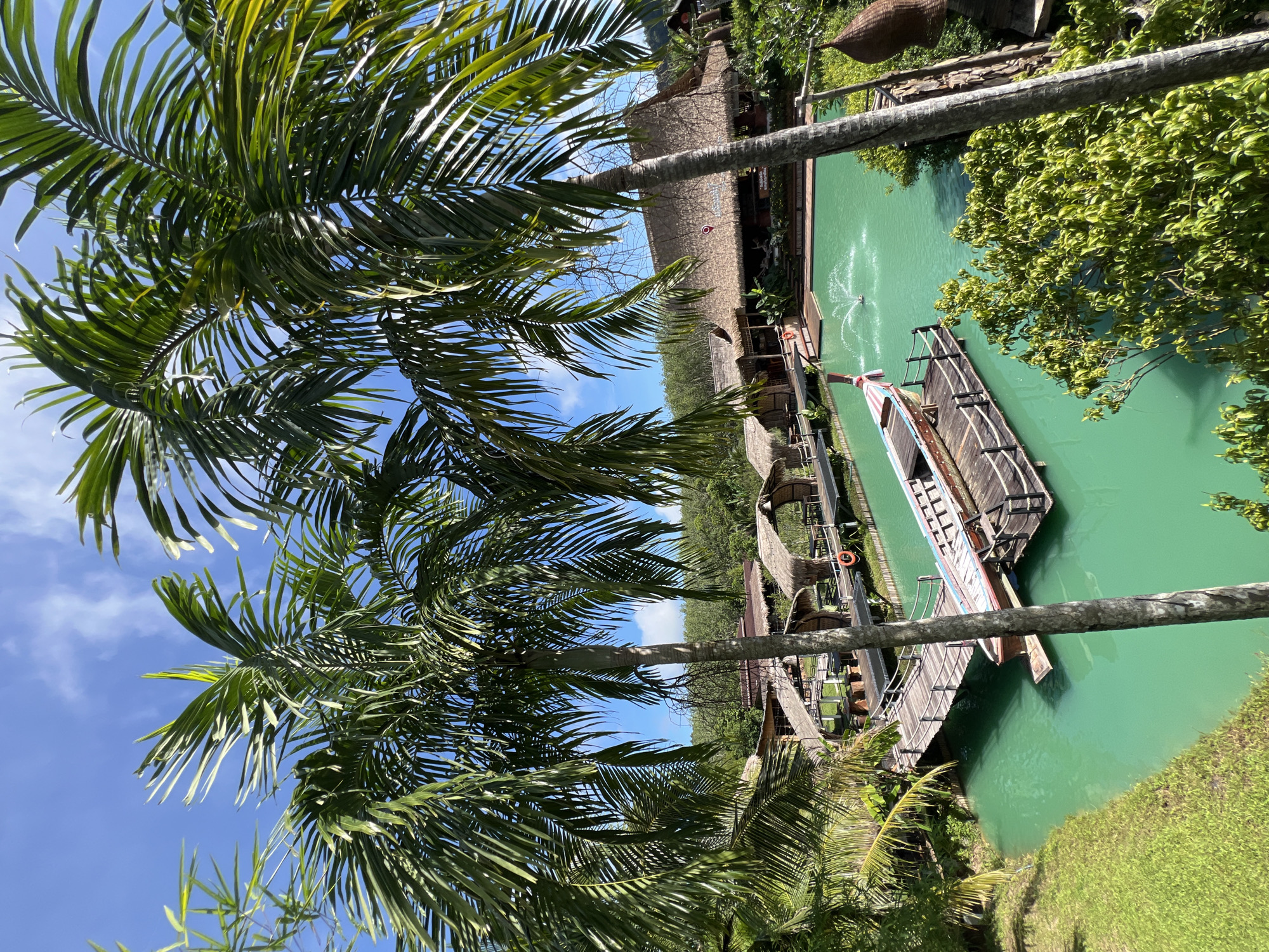 This is a photograph taken from an elevated vantage point in Tambon Pa Kae Bo Hin, Thailand. The scene is dominated by a vibrant turquoise body of water, likely a man-made pond or lagoon. On the water, a longtail boat, a traditional Thai vessel, is moored to a wooden pier. The pier extends from the shore, connecting to several thatched-roof structures nestled among lush greenery. These structures appear to be rustic huts or bungalows, suggestive of a resort or eco-tourism destination. In the background, more thatched-roof buildings are visible, possibly restaurants or communal areas, with their roofs blending into the dense foliage. Two large palm trees frame the foreground of the image, their fronds casting shadows and adding to the tropical ambiance. The sky is a clear blue with a few wispy white clouds. A fountain sprays water in the center of the pond, adding a dynamic element to the otherwise tranquil scene. The overall impression is one of a peaceful, tropical retreat.