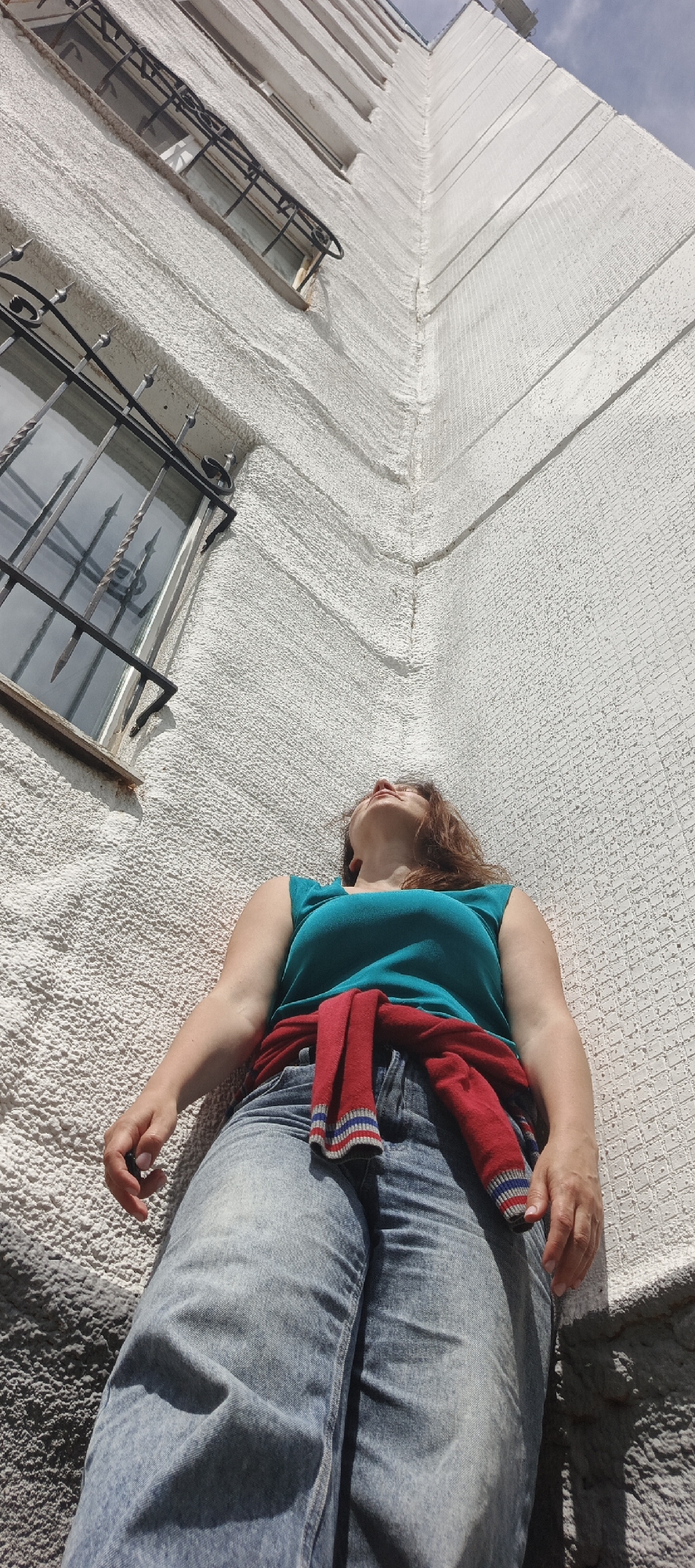 A low-angle, full-body shot captures a woman standing near the corner of a textured white building, her head tilted back and eyes looking up towards the sky. She is wearing a turquoise sleeveless top, light-wash denim jeans, and a red sweater tied around her waist. The building features a rough, stucco-like texture and a window with wrought-iron bars. The scene appears to be outdoors, possibly in a residential area of Chaikovsky, Russia, as indicated by the architectural style and building materials common in the region. The lighting suggests a bright, sunny day, with shadows cast on the building. The woman's pose and upward gaze imply contemplation or admiration of the structure or the sky.