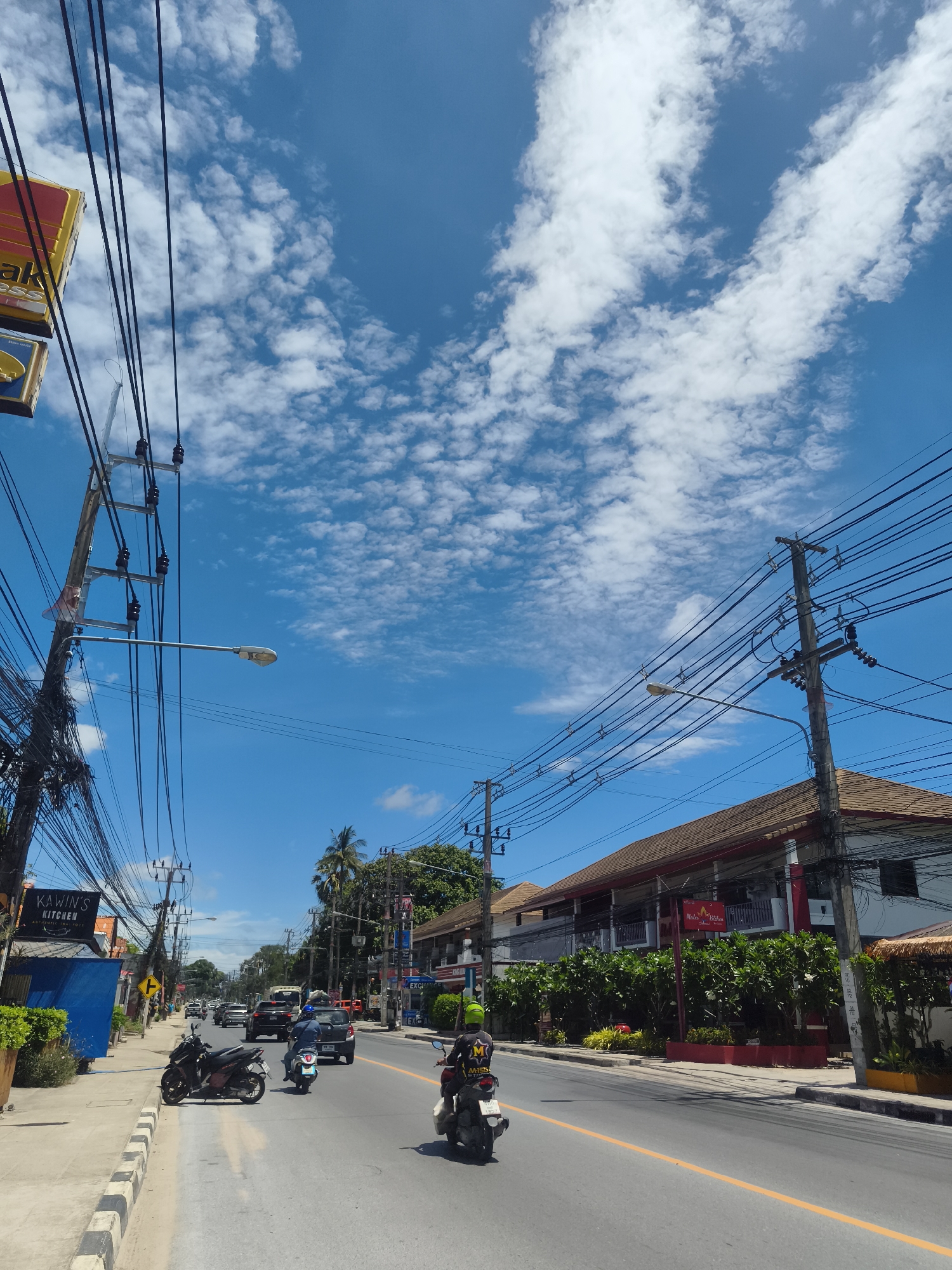 The image shows a street in Baan Bo Phut, Thailand, under a bright blue sky with scattered clouds. The road has two lanes and a yellow line dividing them. Cars and motorbikes are traveling on the road. There is a sidewalk on the left side of the image. Power lines and utility poles run along both sides of the street. Buildings and businesses are visible on either side of the road, including "Kawin's Kitchen" and "Malee Kitchen." The overall scene depicts a typical street scene in a tropical, urban environment.