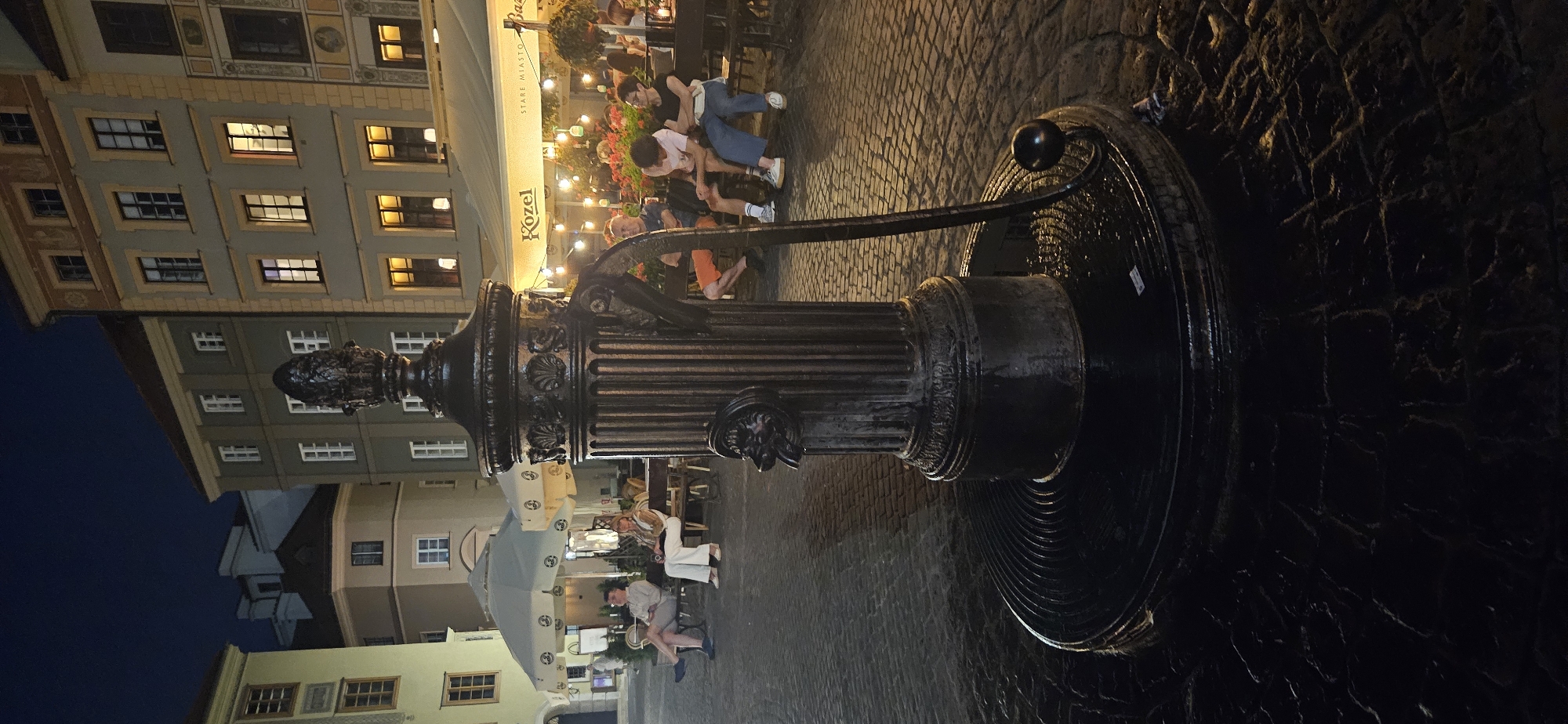 The image depicts an outdoor scene at night, likely in a public square in Warsaw, Poland. A prominent, ornate, dark metal water pump stands in the foreground, its base on a cobblestone surface. The pump is designed with fluted detailing and features a decorative element resembling a lion's head with a spout. A curved metal handle is attached to the pump.
In the background, several people are seated on benches, some at tables, suggesting an outdoor seating area for a restaurant or cafe. The buildings behind them are multi-story structures with illuminated windows, indicating that some of them are occupied or businesses are open. An awning with the word "Közel" is visible, and string lights are hung across the area, illuminating the space. The overall atmosphere is relaxed, with people enjoying the evening. The cobblestone ground appears wet, reflecting the ambient light.