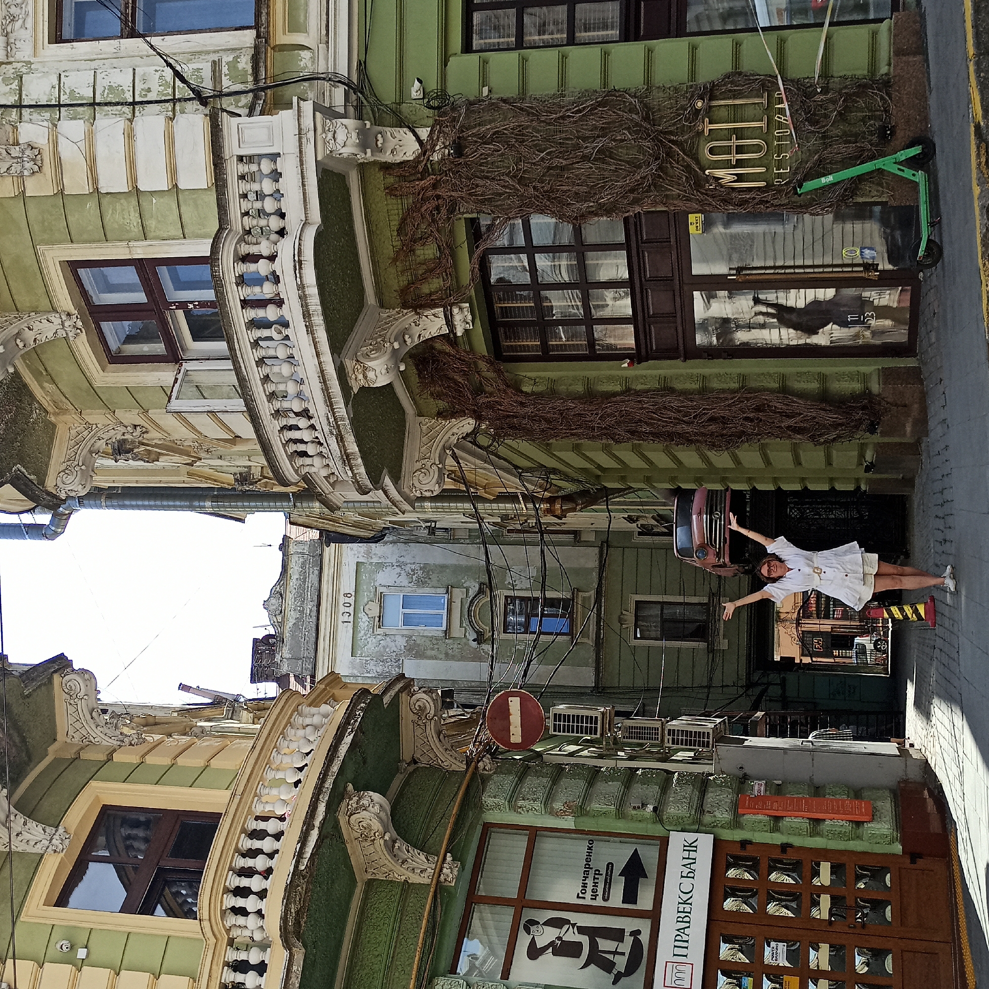 The image captures a street scene in Chernivtsi, Ukraine, specifically the area around the Chernivtsi National University complex, known for its unique architectural blend. In the foreground, a woman dressed in a white outfit, including a short dress and shorts, stands with her arms raised in a celebratory pose. She is positioned on a cobblestone street with a yellow and black striped barrier to her right. Behind her, an old, pink car appears to be suspended in mid-air, attached to the facade of a building.
The architecture is a prominent feature, showcasing two ornate buildings with green facades, cream-colored decorative elements, and balconies with white railings. One building on the left has a window with a sign for "GONCHARENKO CENTER education and culture" and another sign for "PRAVEKS BANK". The building on the right features a business sign that reads "MOTT RESTOBAR" in gold lettering and has a green electric scooter parked in front of it. Several air conditioning units are mounted on the walls, and a prominent tangle of wires runs across the buildings. The overall impression is a vibrant, slightly weathered urban environment with a touch of artistic display.
