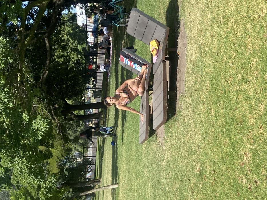 The image captures a sunny daytime scene in a park in Buenos Aires, Argentina. The primary subject is a woman in a multi-colored bikini, smiling directly at the camera. She is leaning on a wooden picnic table, with one arm extended along the table surface and one leg bent and resting on the table bench. A yellow pair of sandals, a pink pair of flip-flops, and a red, white, and blue striped towel are placed on the table next to her.
In the background, several other individuals are visible, enjoying the outdoor setting. Some are sitting on a mat on the grass, while another person is seen with a bicycle. A black metal fence runs horizontally through the mid-ground, separating the foreground activity from further background elements. Large, leafy green trees dominate the left side and the distant background, providing ample shade. The ground throughout the park is covered in vibrant green grass. The overall atmosphere suggests a relaxed, leisurely day in a public park. No discernible text is visible on any objects.