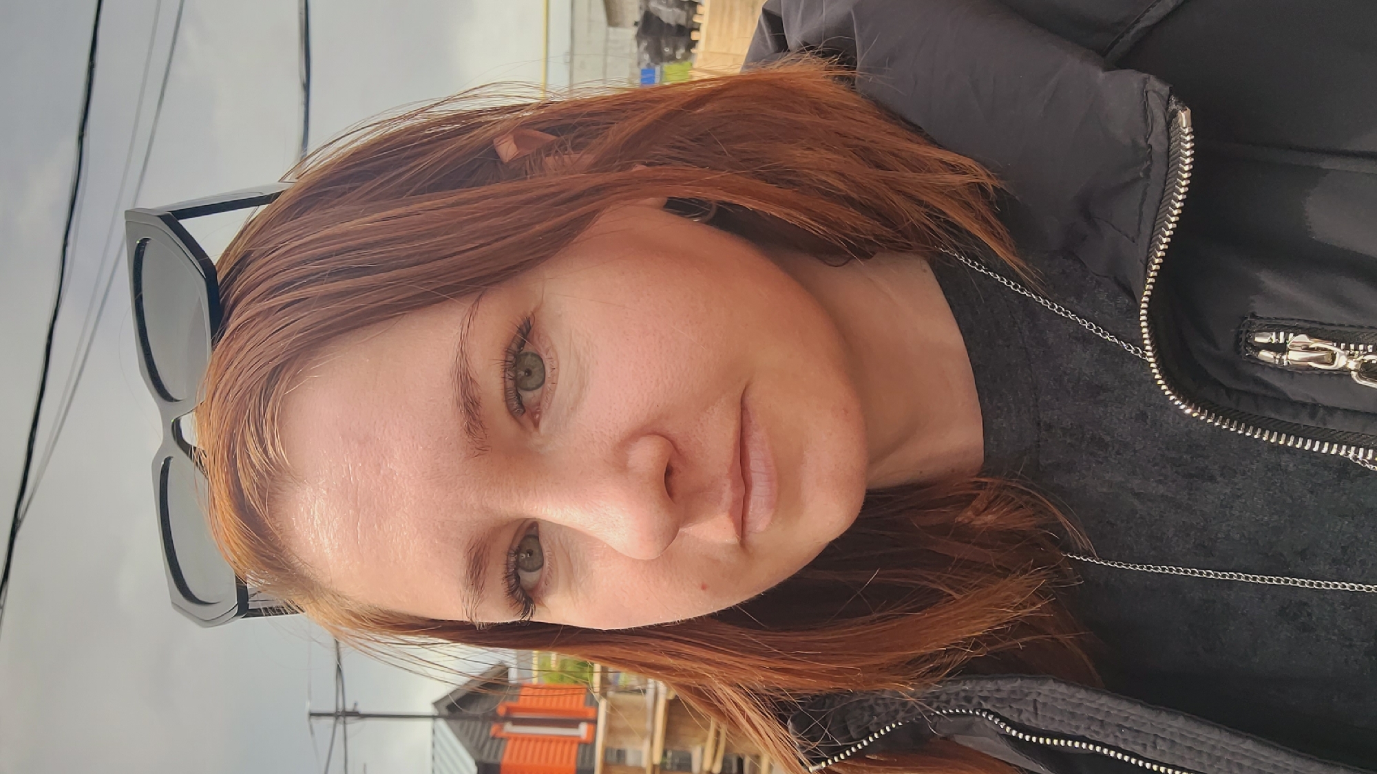 A close-up, selfie-style shot depicts a woman with auburn hair and fair skin. She wears black sunglasses perched on her hair and a black zippered jacket over a dark gray shirt. A silver chain necklace is visible. The background is out of focus, showing an urban environment with power lines and a building with orange accents. The location is Unknown, Unknown.