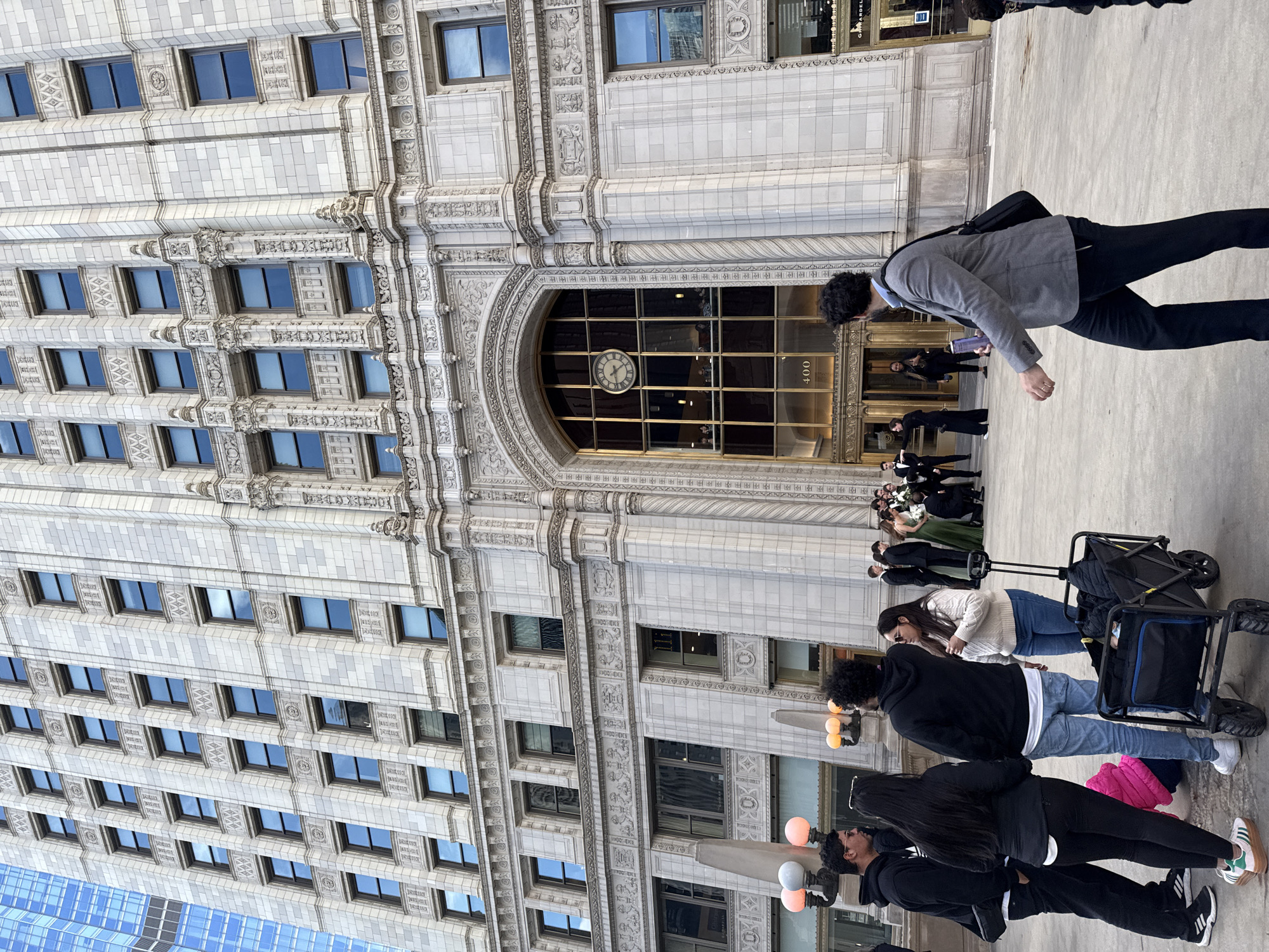The image depicts the exterior of the historic Reliance Building, located at 1 W Washington Street in downtown Chicago. The building's distinctive white terra cotta facade with rows of windows and intricate architectural details are visible. A clock is visible in the entryway along with the address "400." Several individuals are walking in front of the building and standing near the entrance. Some individuals are dressed in formal attire.