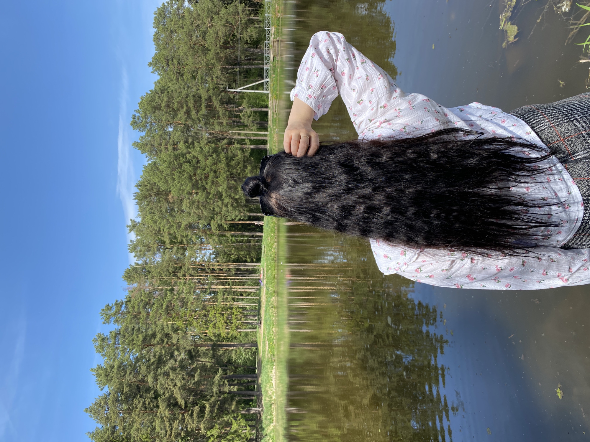 The image depicts a person with long, black hair, wearing a white floral-patterned blouse and grey trousers, lying down near a body of water. The person's head is turned away from the camera, and one hand is touching the hair. The background includes a row of tall, green trees reflected in the water, and a clear blue sky. A portion of a fence structure is visible further back. The location appears to be a lake or pond surrounded by a wooded area, suggestive of a natural or park setting. Chaikovsky, Russia, is not evident in the picture.