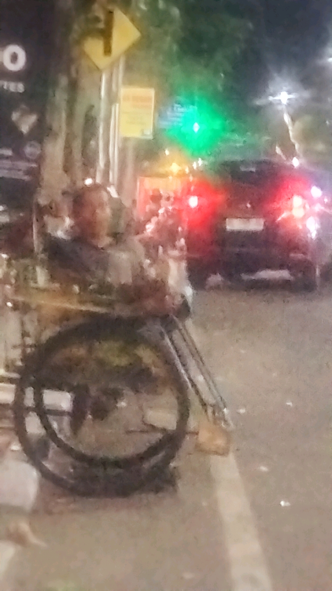 A low-light, slightly blurry medium shot shows a person in a wheelchair on a sidewalk. The person is facing slightly to the left and appears to be wearing a light-colored shirt. The wheelchair has large spoked wheels. Behind the wheelchair, a street is visible with traffic lights showing green, and a dark-colored car with its red taillights illuminated. A yellow diamond-shaped road sign with a black arrow pointing to the right is mounted on a pole to the left of the traffic lights. A smaller yellow sign is below the diamond sign. The scene is set at night, suggesting a street in Bojonegoro, Indonesia, likely near an intersection with traffic control.