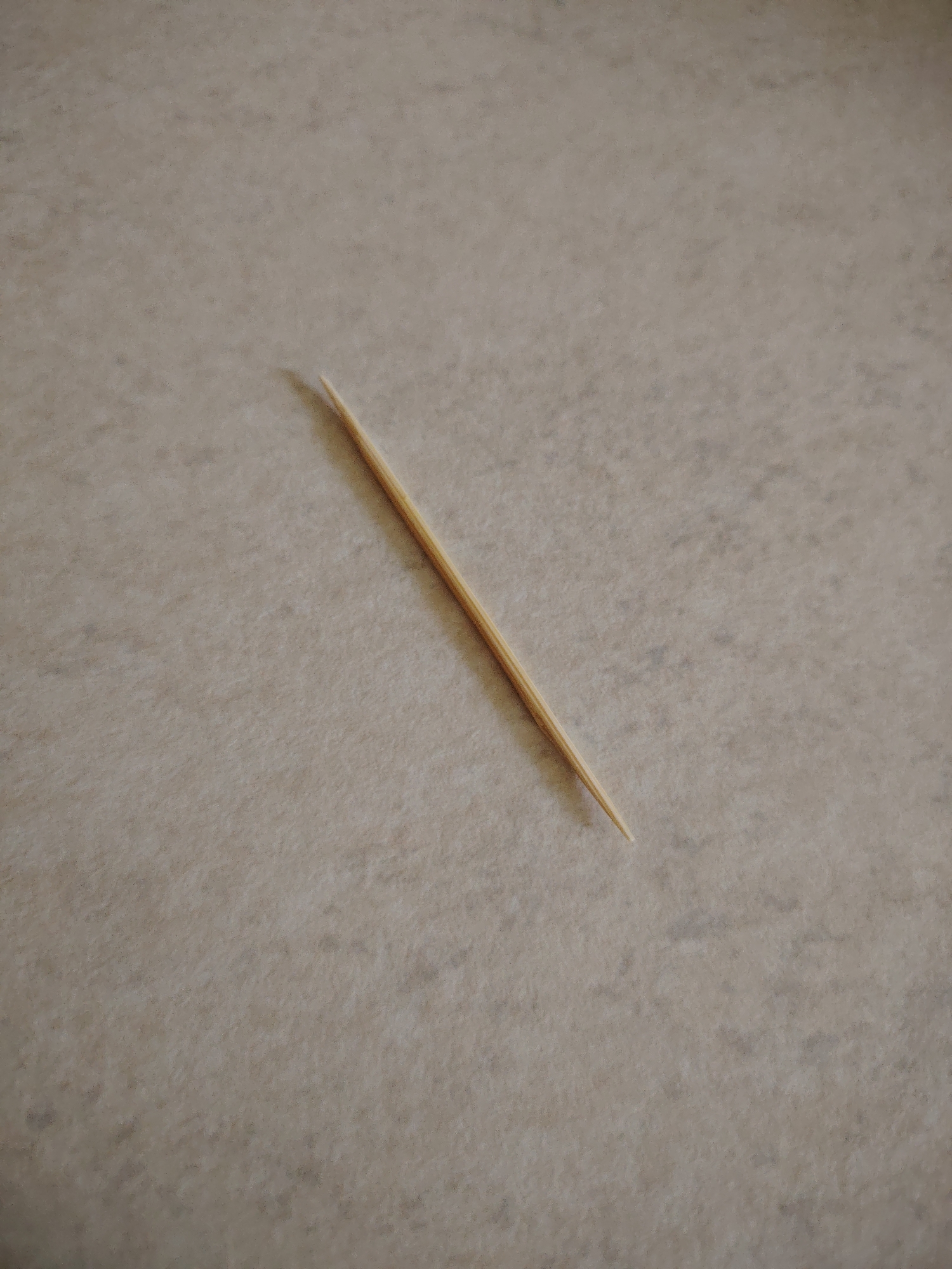 A single wooden toothpick lies diagonally on a textured, light-colored surface. The toothpick is positioned from the lower right towards the upper left of the frame. The surface appears to be a countertop or a similar material with a subtle speckled pattern. The lighting casts a soft shadow from the toothpick, indicating a light source from above and slightly to the left. No people or other objects are visible in the image. The scene is static and focused on the solitary toothpick.