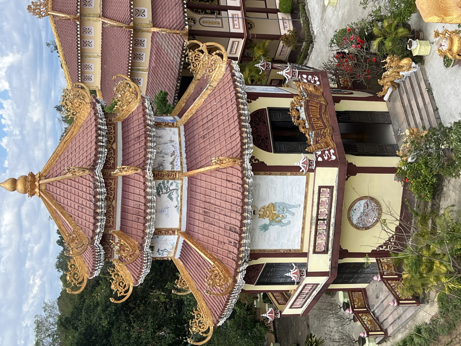An octagonal, multi-tiered pagoda, identified by signage as "THÁP QUÁN ÂM" (Thap Quan Am), stands in the city of Đăk Mar, Vietnam. The structure features terracotta-tiled roofs with elaborate golden ornate decorations, including stylized dragon motifs on the eaves and a prominent golden finial at the apex. Its walls are painted cream-yellow with dark brown trim and are adorned with various murals and bas-reliefs depicting figures, landscapes, and circular medallions. The pagoda has multiple levels, each with recessed windows or doors and, on the lower visible tier, a balcony with small red-roofed lantern-like elements on the railings. Stone steps lead to several entrances. In the foreground and surrounding areas, green foliage, trees, potted plants, and flowering bushes are visible. Golden guardian statues flank an entrance on the lower level, and an ornate golden incense burner is partially visible in the lower right. A portion of another building with a similar architectural style, including a tiled roof and cream-colored walls, is visible to the right. The sky overhead is partly cloudy blue. No individuals are present in the scene.