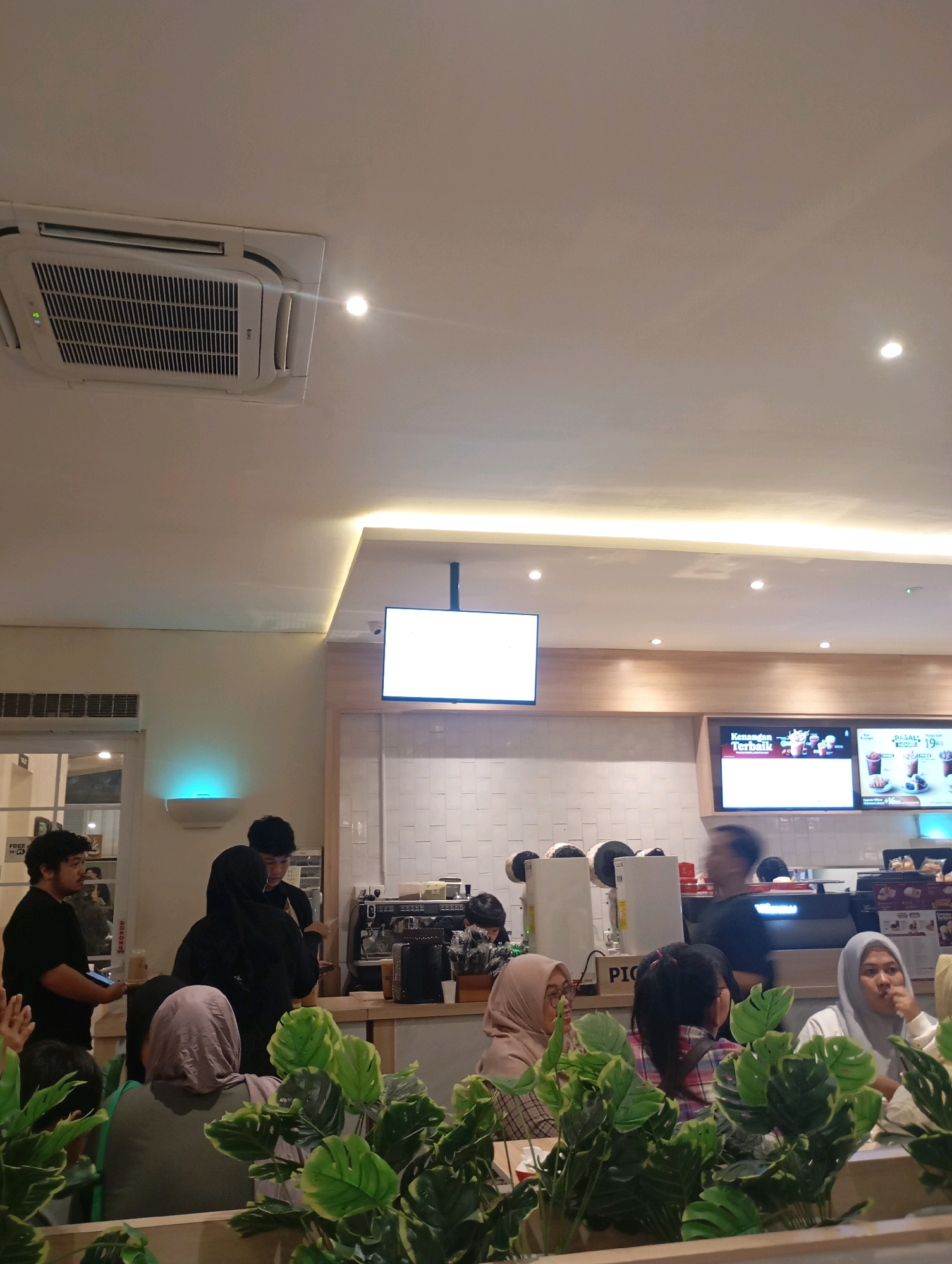 The image depicts the interior of a busy cafe, possibly a coffee shop or fast-food establishment, with patrons seated and staff behind a counter. In the foreground, lush green artificial plants create a partial barrier between the viewer and the seating area. Several individuals are visible, many wearing headscarves, engaged in conversation or looking towards the counter. Behind them, staff members attend to the counter area, where coffee machines and a menu display are present. A large screen displays advertisements or menu items. The ceiling features a prominent air conditioning unit and recessed lighting. The overall ambiance suggests a casual dining or social gathering space. The scene is consistent with a typical cafe found in urban areas of Tangerang, Indonesia, a city known for its numerous commercial and dining establishments.