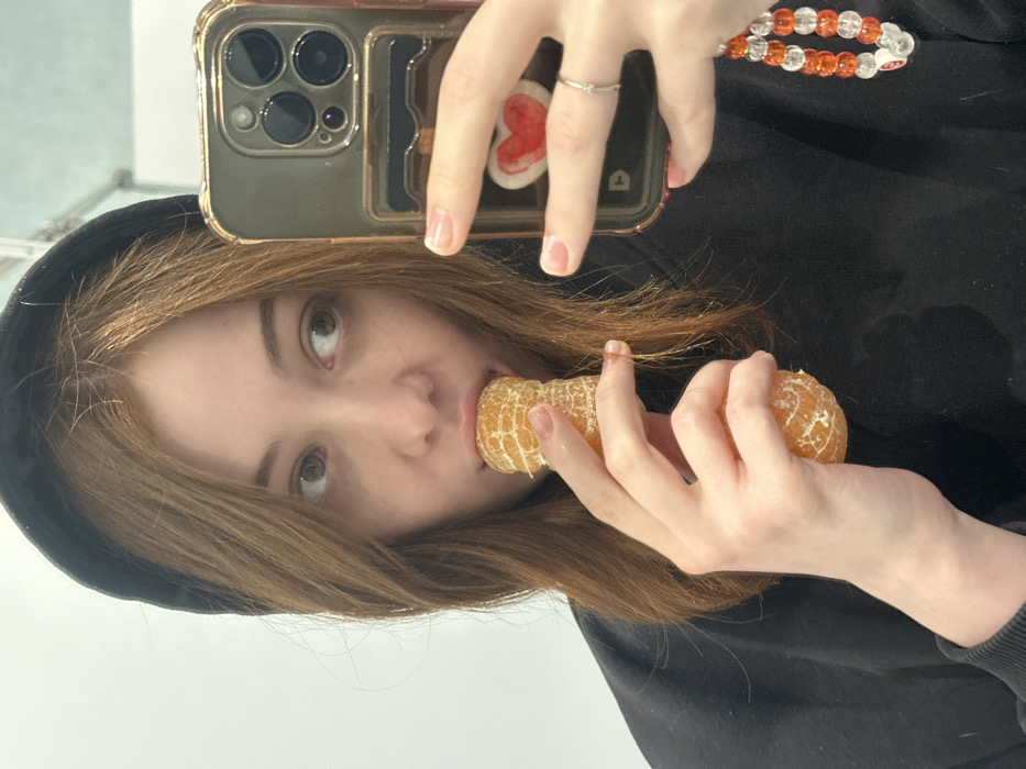 A person in a black hoodie is taking a selfie while eating a peeled orange. The person's left hand holds the phone, which has a clear case with a heart-shaped design and a beaded strap. The right hand holds the orange close to the person's mouth, and the person is looking directly at the camera with a slight tilt of the head. The person's hair is light brown and falls loosely around the face. The background is a plain white wall.