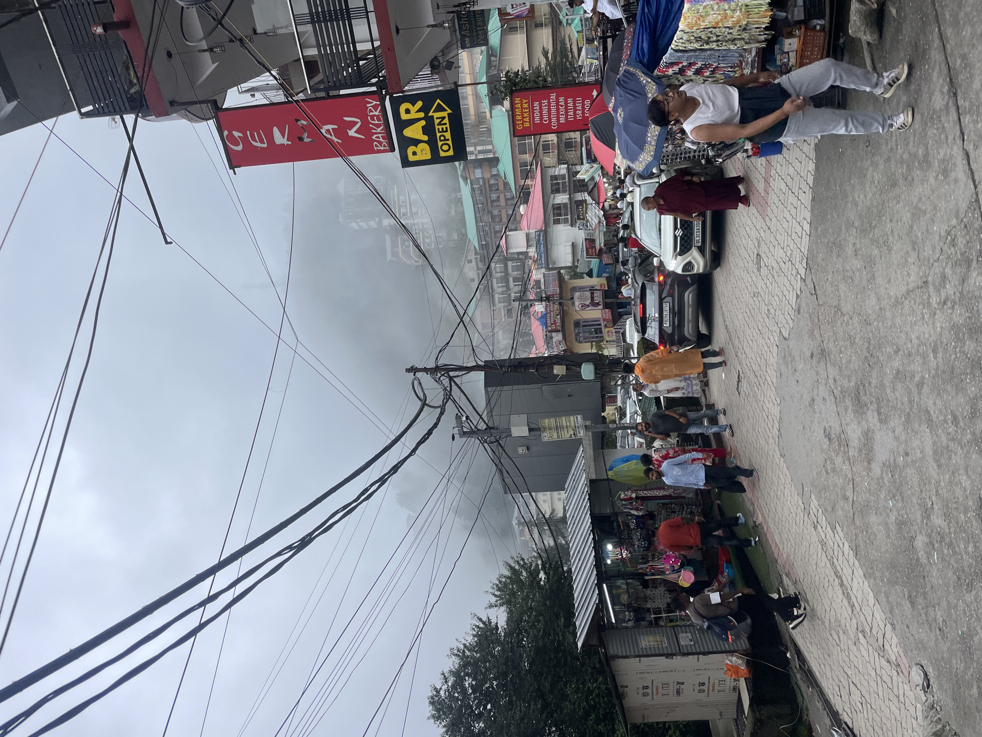 This image depicts a bustling street scene in Bhagsu, India, shrouded in a light mist. The foreground shows a paved walkway with several individuals. To the left, a small market stall is set up with various items displayed for sale, and people are browsing. A group of men, one in a red shirt and another in a blue shirt, walk along the walkway. Further down, a man in a maroon robe is visible walking beside a white SUV. In the midground, a busy street is lined with buildings and shops. A prominent red banner with white text reading "GERMAN BAKERY" is attached to a building, and below it, a yellow sign with black text says "BAR OPEN". More people are seen walking and interacting in this area. The background reveals multi-story buildings with colorful roofs, typical of Indian architecture, ascending a hillside. The overall atmosphere suggests a vibrant commercial area with a steady flow of pedestrian and vehicle traffic, all under a hazy sky.