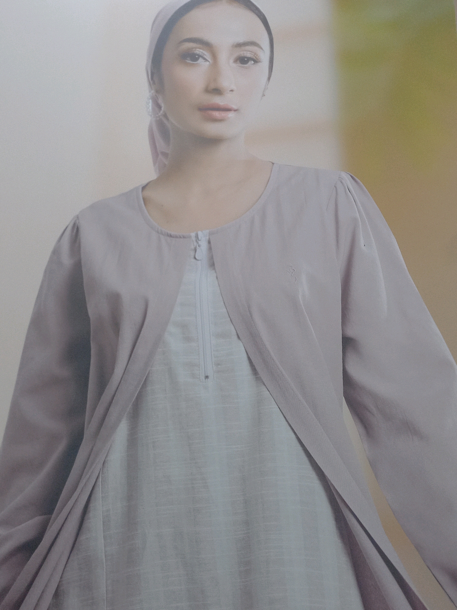 The image is a portrait of a young woman wearing a light purple headscarf and a light purple outer garment over a pale blue inner garment. The outer garment has a rounded neckline and long sleeves with puffed shoulders. The inner garment has a zipper down the front. The woman is looking directly at the camera with a neutral expression. The background is a soft, blurred gradient of beige and pale green, suggesting an indoor studio setting. There is a subtle logo or emblem on the right breast area of the outer garment. The image is a medium shot, focusing on the woman's upper body and face. The scene is well-lit with soft, diffused light. There is no identifiable geographic information within the image itself to indicate a specific location in Tunggorono, Indonesia.