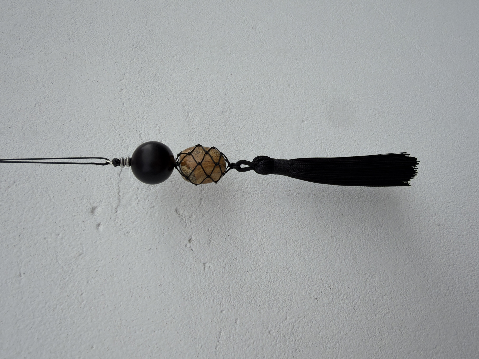 A close-up, eye-level, medium shot captures a hanging decoration against a textured, white, concrete wall. The decoration, suspended by a thin black string, features a dark, spherical bead at the top, followed by a lighter, rough-textured stone encased in a fine, dark netting. Below the stone, a black tassel with tightly bound threads hangs loosely. The lighting is even, with no strong shadows, suggesting an indoor or overcast outdoor setting. The overall aesthetic is minimalist and monochromatic, with the white wall providing a neutral backdrop that emphasizes the details of the hanging ornament. The image does not contain any identifiable individuals or specific urban features that would allow for a precise location within Khimki, Russia.