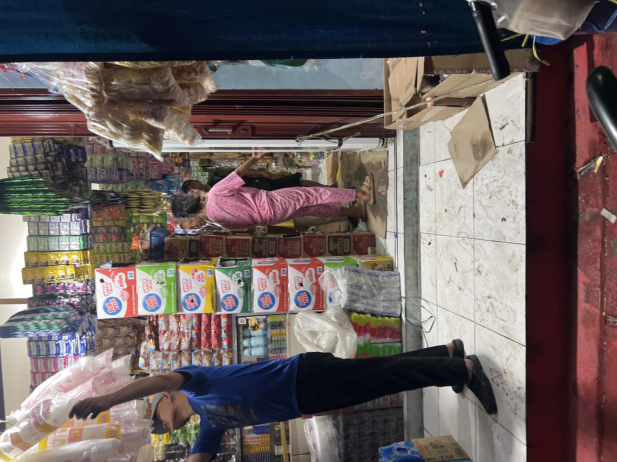 A medium shot captures the interior of a cluttered convenience store in Citeureup, Indonesia, on a dimly lit evening. In the foreground, a person wearing a blue shirt and black pants, with a cap on their head, reaches up to adjust a stack of white disposable cups hanging from the ceiling. To their right, the store is filled with shelves stocked with various packaged goods, including boxes of instant noodles with colorful branding and bags of snacks.
Further into the store, two individuals, one wearing a pink patterned outfit and the other in a dark shirt and pants, are standing near a closed metal roll-up door. The person in pink is looking towards the front of the store, while the person in dark clothing appears to be facing away. The floor is tiled with a white and grey pattern, scattered with debris and cardboard. The overall scene depicts a typical small retail environment with goods stacked high and displays of merchandise.