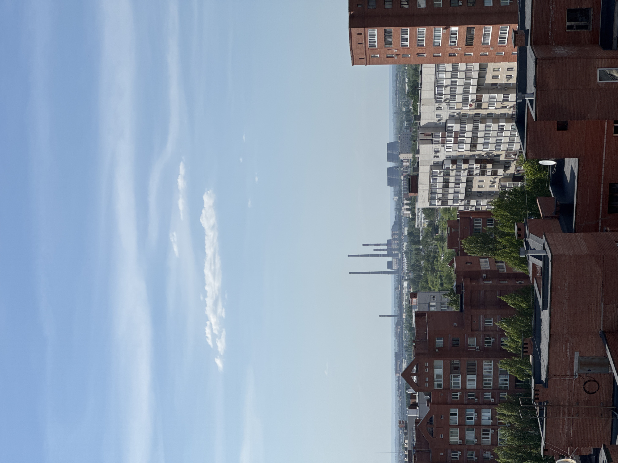 Here is a description of the image:
The image shows a cityscape, likely taken from a high vantage point. On the right side of the frame, there are rows of brick buildings with numerous windows, some of which have trees planted in front. The style of architecture suggests a residential area. In the distance, there are taller structures and smokestacks that hint at an industrial presence in the city. On the left side, the sky occupies a significant portion of the frame, with streaks of clouds against a light blue background. The sky is clear and free of objects. The image looks as though it was taken on a sunny day.