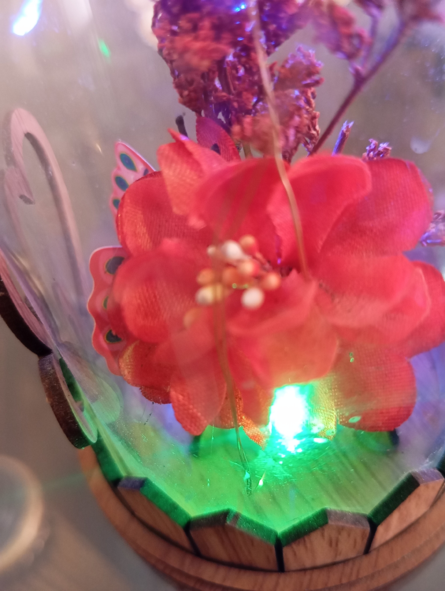 A red artificial flower with small white beads at its center is encased in a glass dome. The flower is partially obscured by the dome's curvature and a green light illuminates the bottom of the dome, casting a green glow onto the flower's petals and the surrounding area. A wooden base with geometric cutouts supports the glass dome. The scene is lit with a combination of purple and white lights, suggesting a decorative or ambient lighting setup.