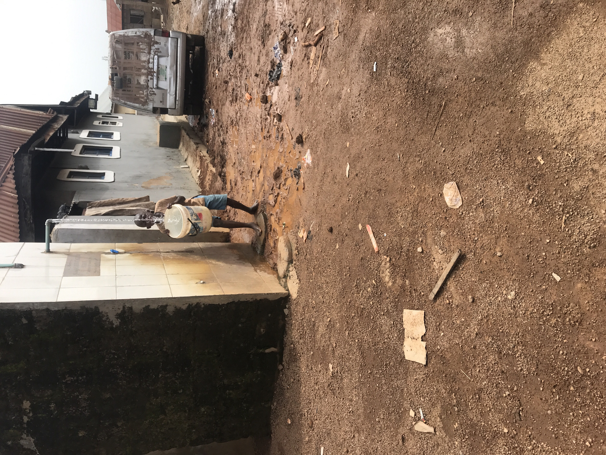 This image captures an outdoor scene in what appears to be a residential or semi-urban area, consistent with the Ilorin, Nigeria context. The central focus is an individual, likely male, standing on a tiled platform, collecting water from a tap into a large white plastic bucket. Water is actively flowing from the tap into the bucket.
To the left of the person is a grey building with a rusty corrugated metal roof and several small, rectangular windows with white frames, some containing blue panes. The base of the tiled platform is covered in dark moss or algae. The ground extending from the platform to the right is unpaved, consisting of reddish-brown dirt and significant patches of mud, indicating wet conditions or recent rain. Scattered across this muddy ground are various pieces of litter, including plastic, cardboard, and small sticks.
Further into the background, to the right of the building, a light-colored minivan or utility vehicle is parked facing away from the viewer. Its rear shows signs of wear or repair with a brown, possibly patched, appearance. The sky is overcast, suggesting daytime with diffused light. No clear text is legible on the bucket, vehicle, or surrounding structures. The scene depicts a routine activity of water collection in a setting characterized by simple infrastructure and natural ground.